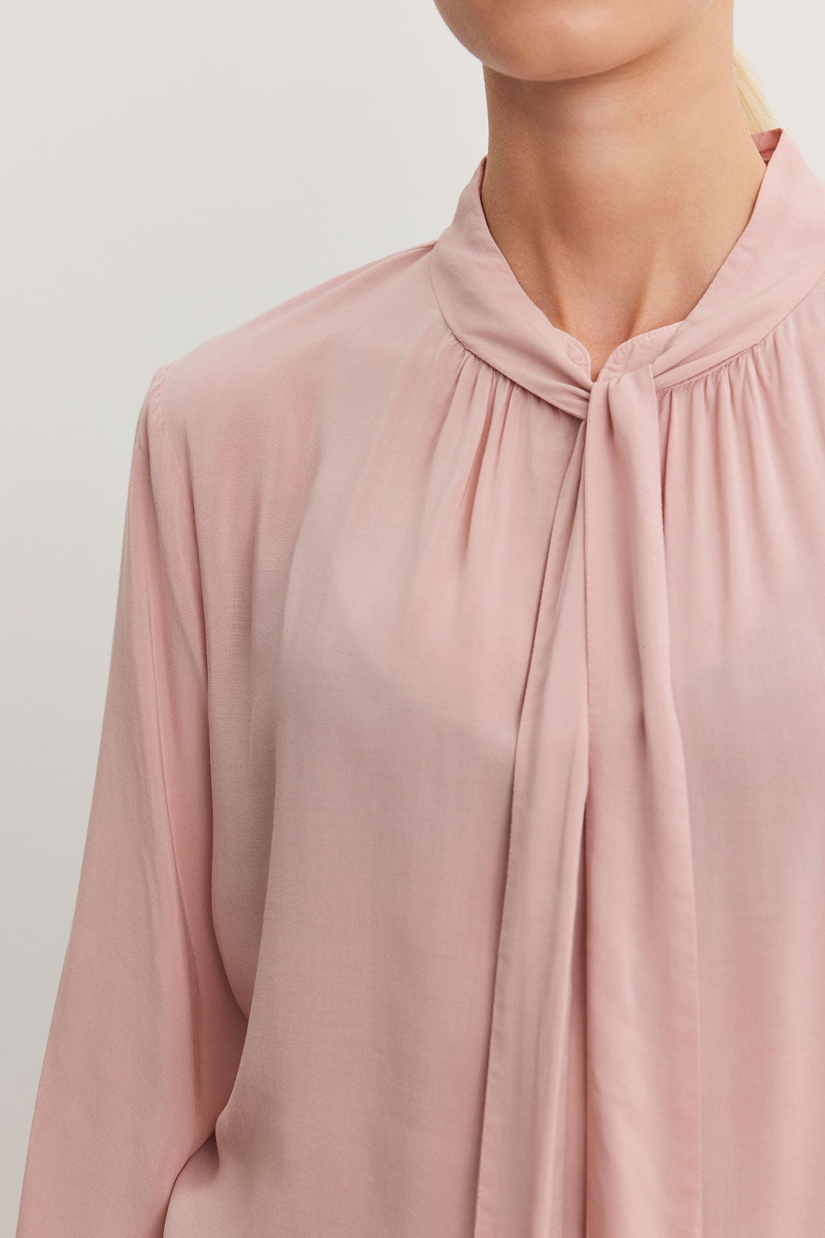 A person is wearing the Gabrielle Top from Velvet by Graham & Spencer, crafted from rayon challis in a light pink hue. It features a gathered neckline with a scarf tie detail and offers a relaxed fit, set against a neutral background.-38301353509057