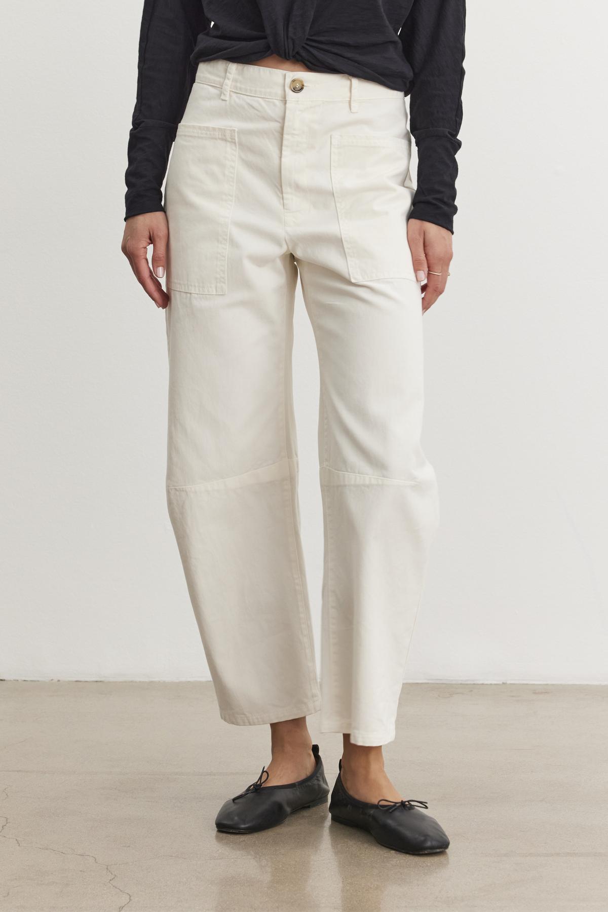   A person stands on a polished concrete floor, wearing Velvet by Graham & Spencer's cream BRYLIE SANDED TWILL UTILITY PANT and a black long-sleeve top. Black ballet flats complete the casual look. 