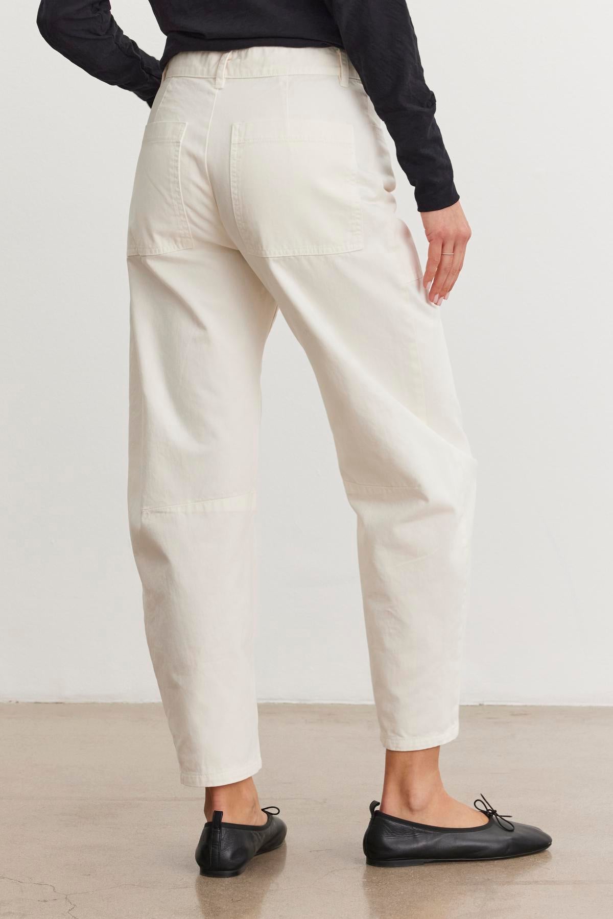   A person wearing Velvet by Graham & Spencer's Brylie Sanded Twill Utility Pant with patch pockets and black flats is viewed from behind, standing on a concrete floor. 