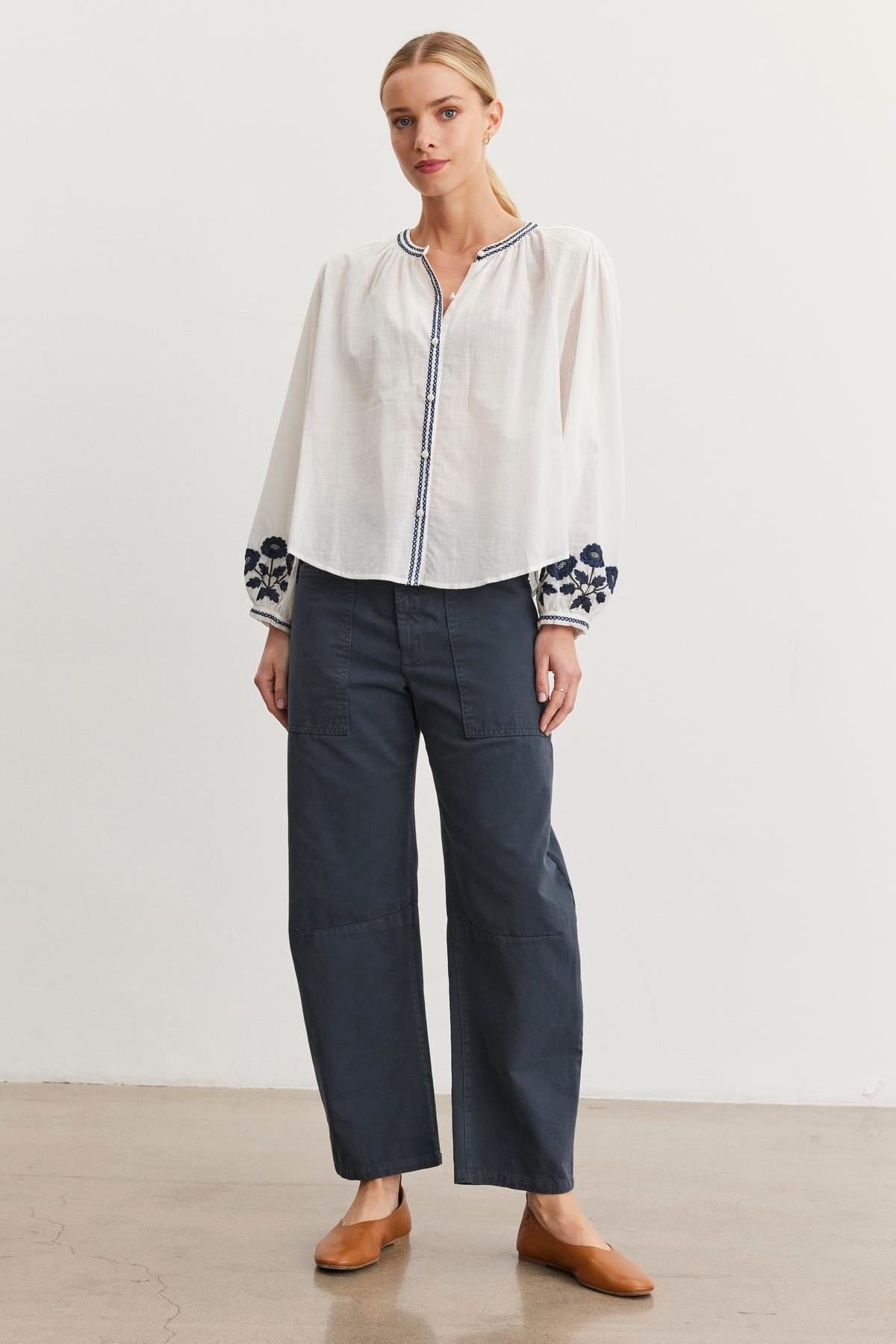 A person stands against a plain white background, wearing a white blouse with floral sleeves, Velvet by Graham & Spencer's dark BRYLIE SANDED TWILL UTILITY PANT featuring patch pockets, and brown shoes.-38512693182657