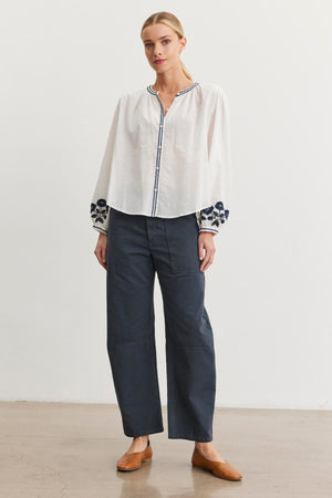 A person stands against a plain white background, wearing a white blouse with floral sleeves, Velvet by Graham & Spencer's dark BRYLIE SANDED TWILL UTILITY PANT featuring patch pockets, and brown shoes.