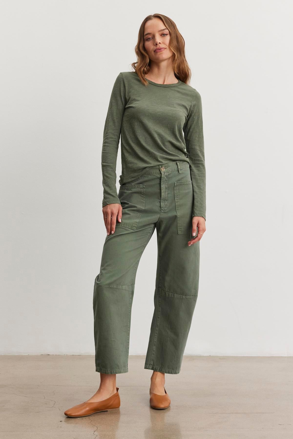   A person stands against a white background wearing the LIZZIE TEE, a long-sleeve green top by Velvet by Graham & Spencer, with matching green pants and brown flats. 