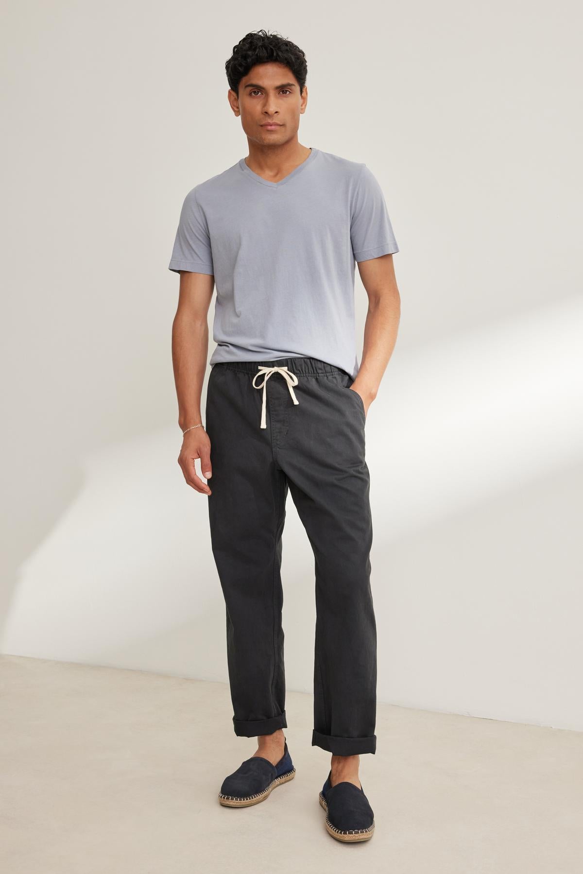   A person stands against a plain background dressed in light gray T-shirt, Velvet by Graham & Spencer's dark BRANSON PANT with an elastic waist and drawstring, paired with casual shoes. 