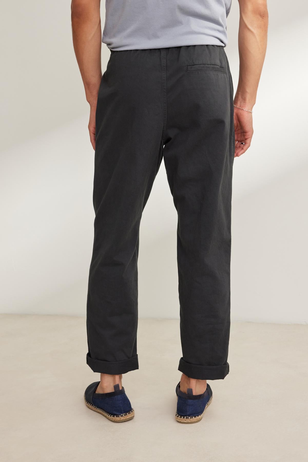   A person in a gray T-shirt and blue shoes stands on a light-colored floor with their back to the camera, wearing Velvet by Graham & Spencer's BRANSON PANT. These dark pants feature an elastic drawstring waist and straight leg design, effortlessly blending comfort with style. 