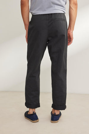 A person in a gray T-shirt and blue shoes stands on a light-colored floor with their back to the camera, wearing Velvet by Graham & Spencer's BRANSON PANT. These dark pants feature an elastic drawstring waist and straight leg design, effortlessly blending comfort with style.