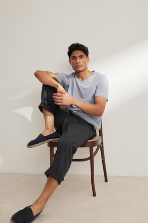 A person sits comfortably, wearing a light gray t-shirt paired with Velvet by Graham & Spencer's BRANSON PANT in dark cotton sanded twill. The elastic drawstring waist ensures a relaxed fit, while their slip-on shoes enhance the effortless style against a plain backdrop.