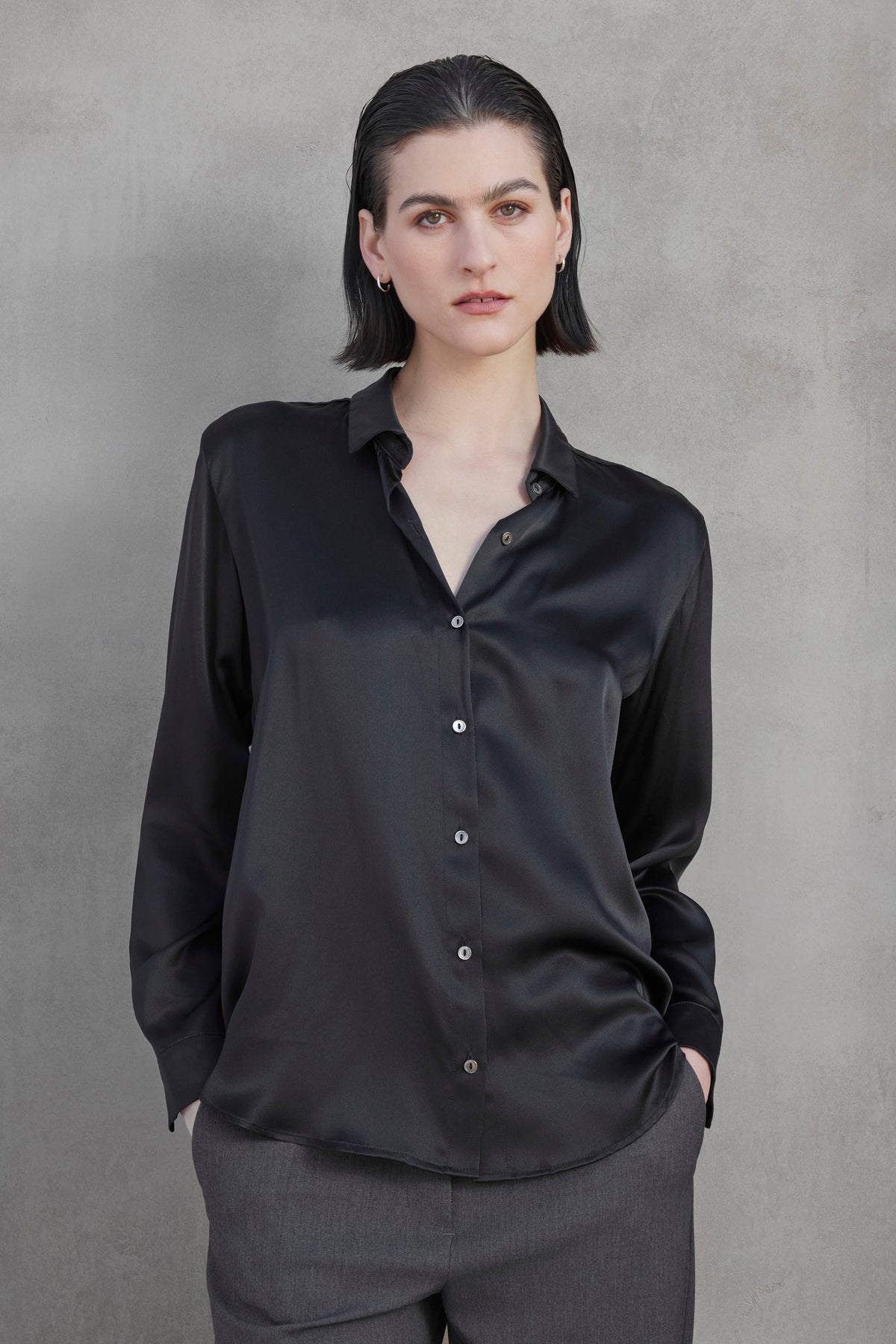   A woman with short dark hair, wearing the Velvet by Jenny Graham CALABASAS SILK CHARMEUSE BLOUSE in black and gray pants, stands against a gray background with her hands in her pockets. 