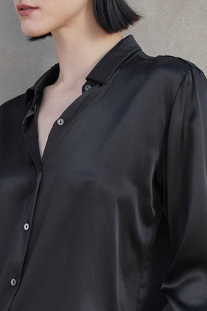 Close-up of the upper body of a person wearing the CALABASAS SILK CHARMEUSE BLOUSE by Velvet by Jenny Graham, featuring a partially open collar, standing against a light brown background.