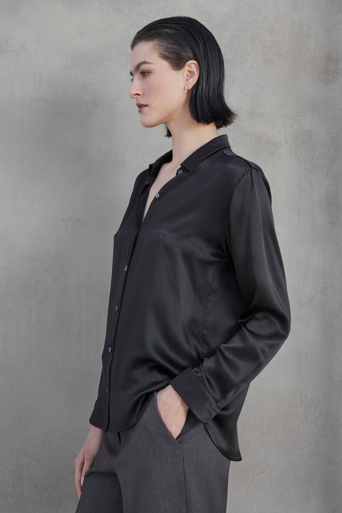   A person with short dark hair wearing the CALABASAS SILK CHARMEUSE BLOUSE from Velvet by Jenny Graham and grey trousers stands against a plain grey background, looking to the side. 