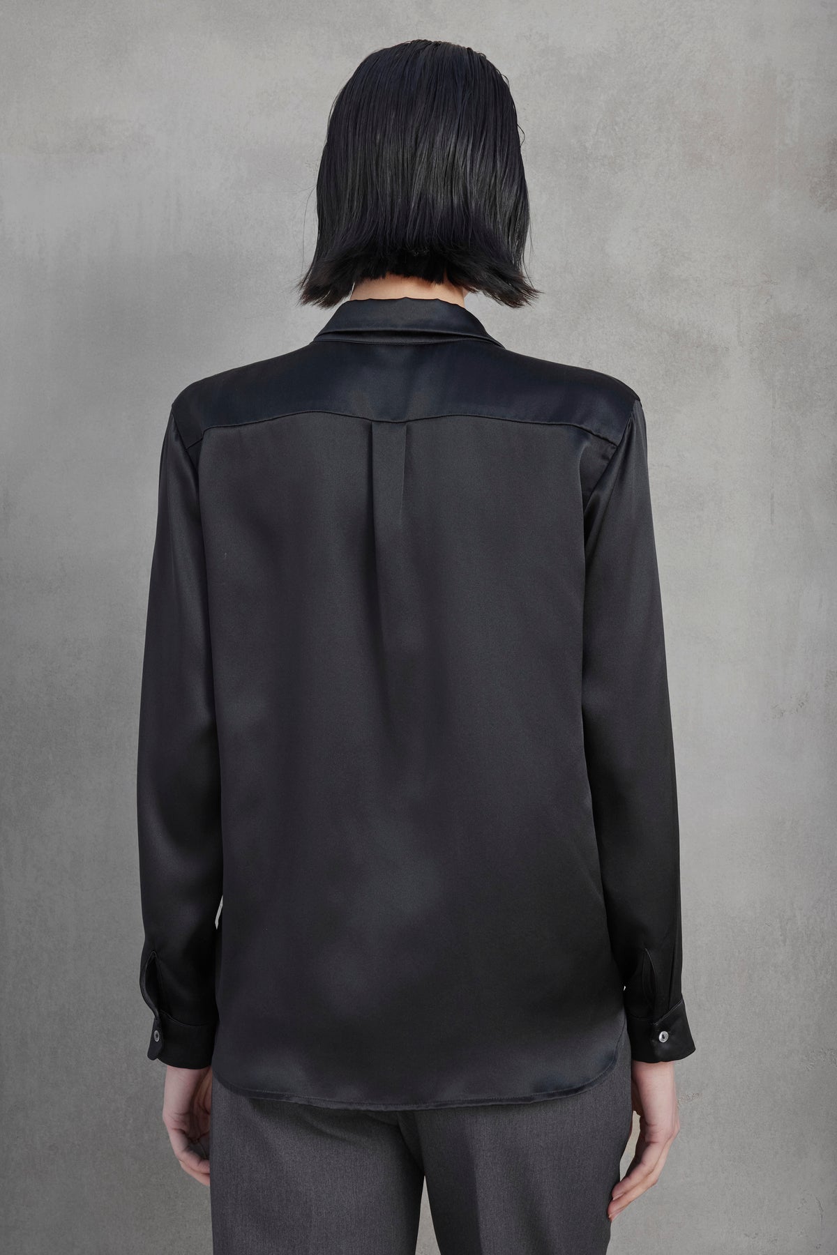   A person with shoulder-length black hair is seen from the back, wearing a CALABASAS SILK CHARMEUSE BLOUSE by Velvet by Jenny Graham and gray pants, standing against a gray wall. 