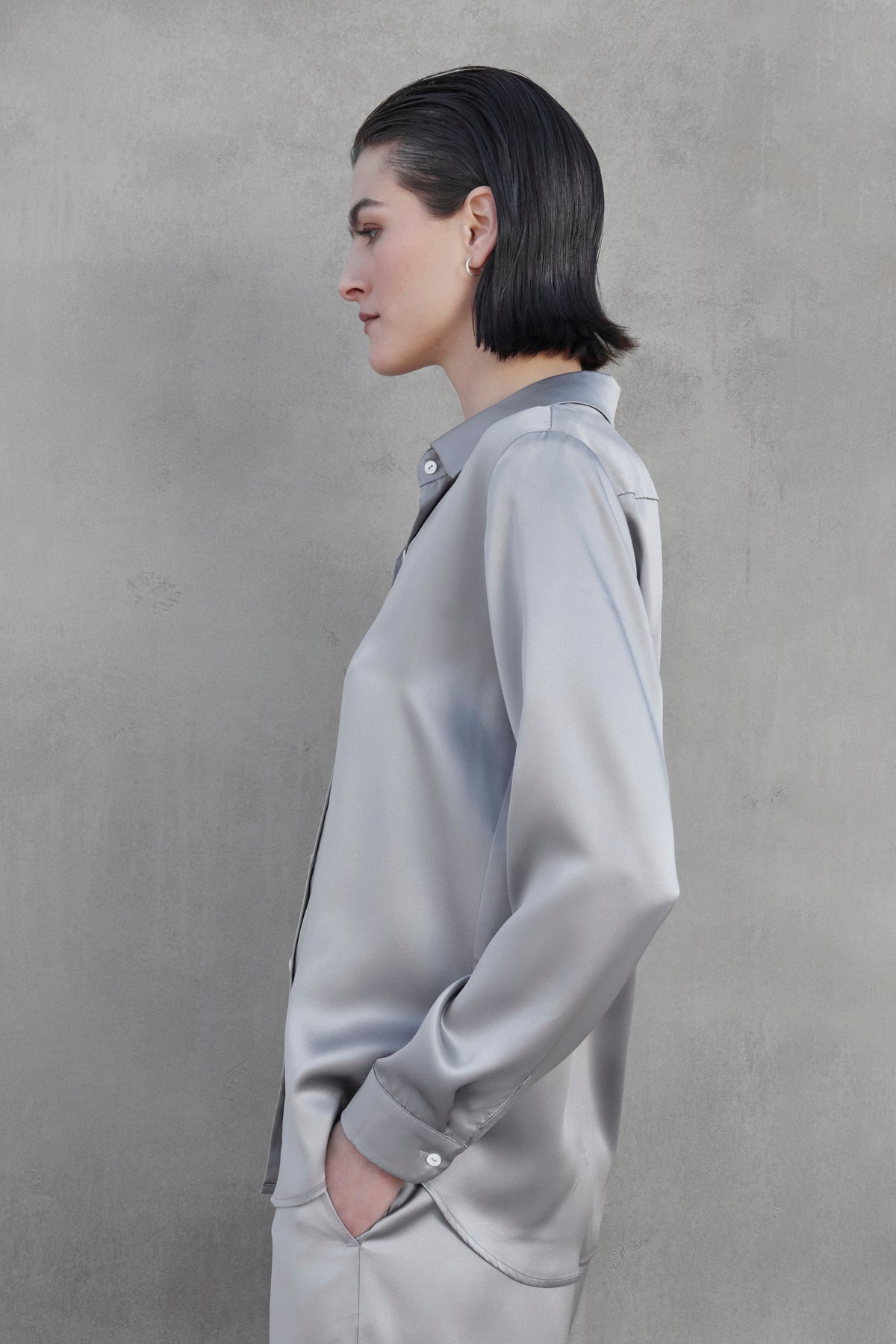   A person with short black hair stands in profile against a gray background, wearing the CALABASAS SILK CHARMEUSE BLOUSE by Velvet by Jenny Graham and matching pants. 