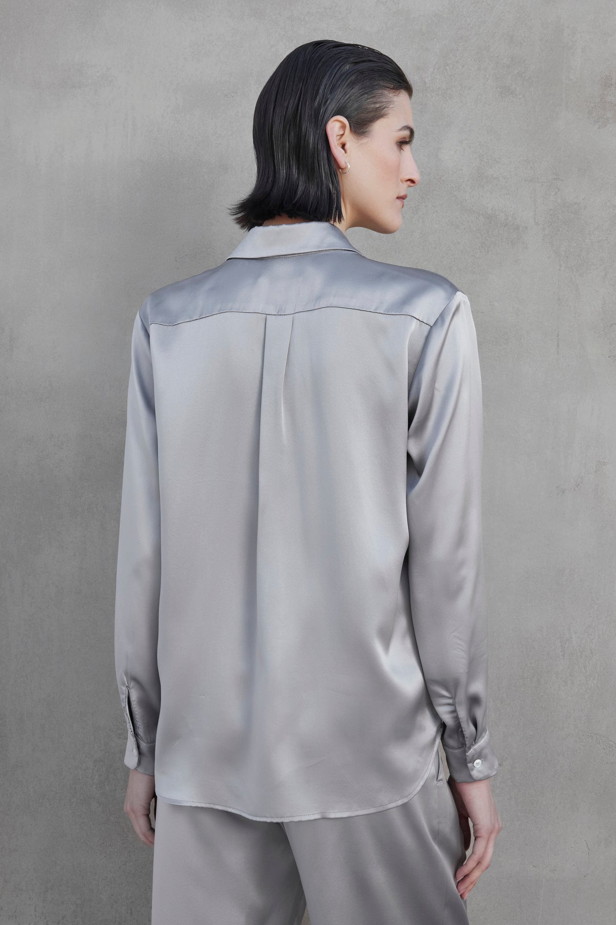   A person in a silver CALABASAS SILK CHARMEUSE BLOUSE by Velvet by Jenny Graham with matching pants faces away, showcasing the garment's back details against a grey background. 