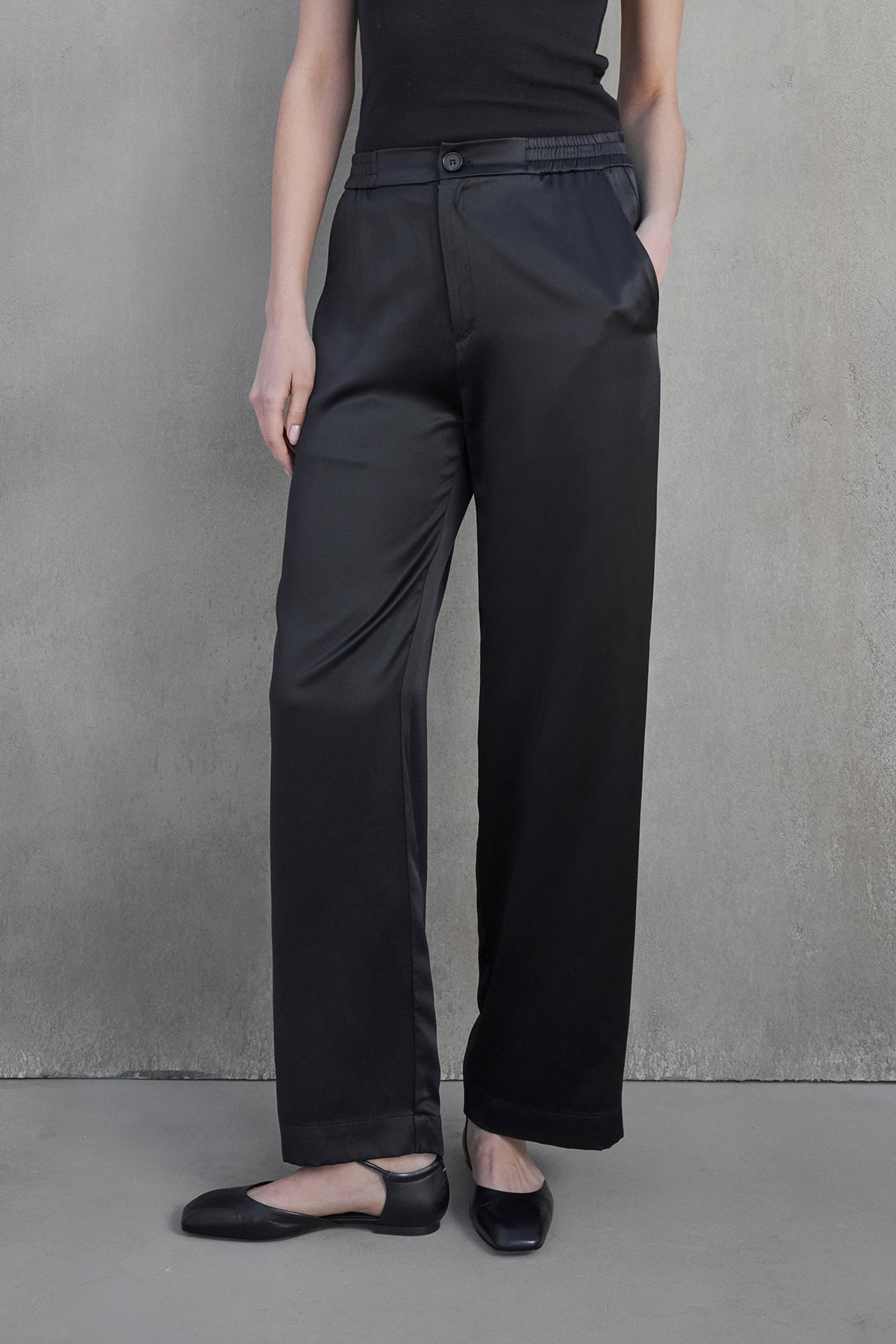   A person wearing a sleeveless black top and the MODESTO SILK CHARMEUSE PANT in black by Velvet by Jenny Graham stands against a plain background, with one arm visible and hands not shown. Black flats are worn on their feet. 