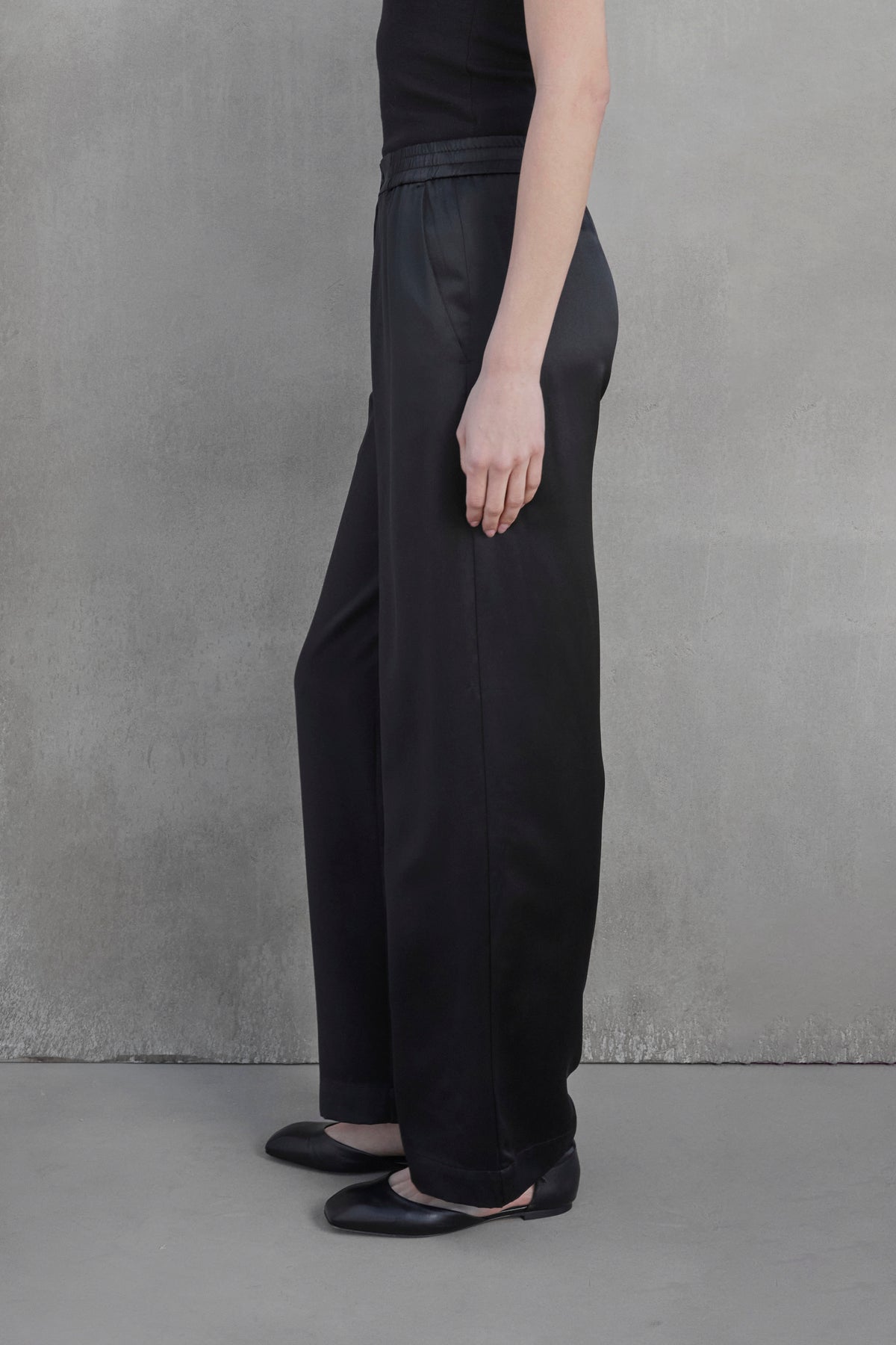   Person standing sideways wearing Velvet by Jenny Graham's MODESTO SILK CHARMEUSE PANT in black, a black top, and black shoes against a gray concrete wall background. 