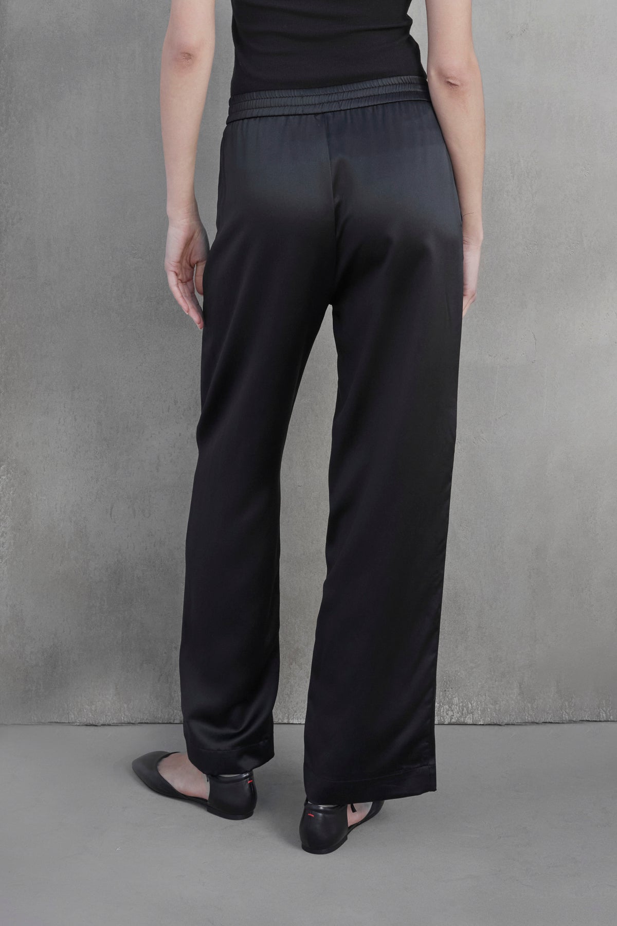   Person standing with their back to the camera, wearing black MODESTO SILK CHARMEUSE PANT by Velvet by Jenny Graham and black flats against a plain, light-colored wall. 