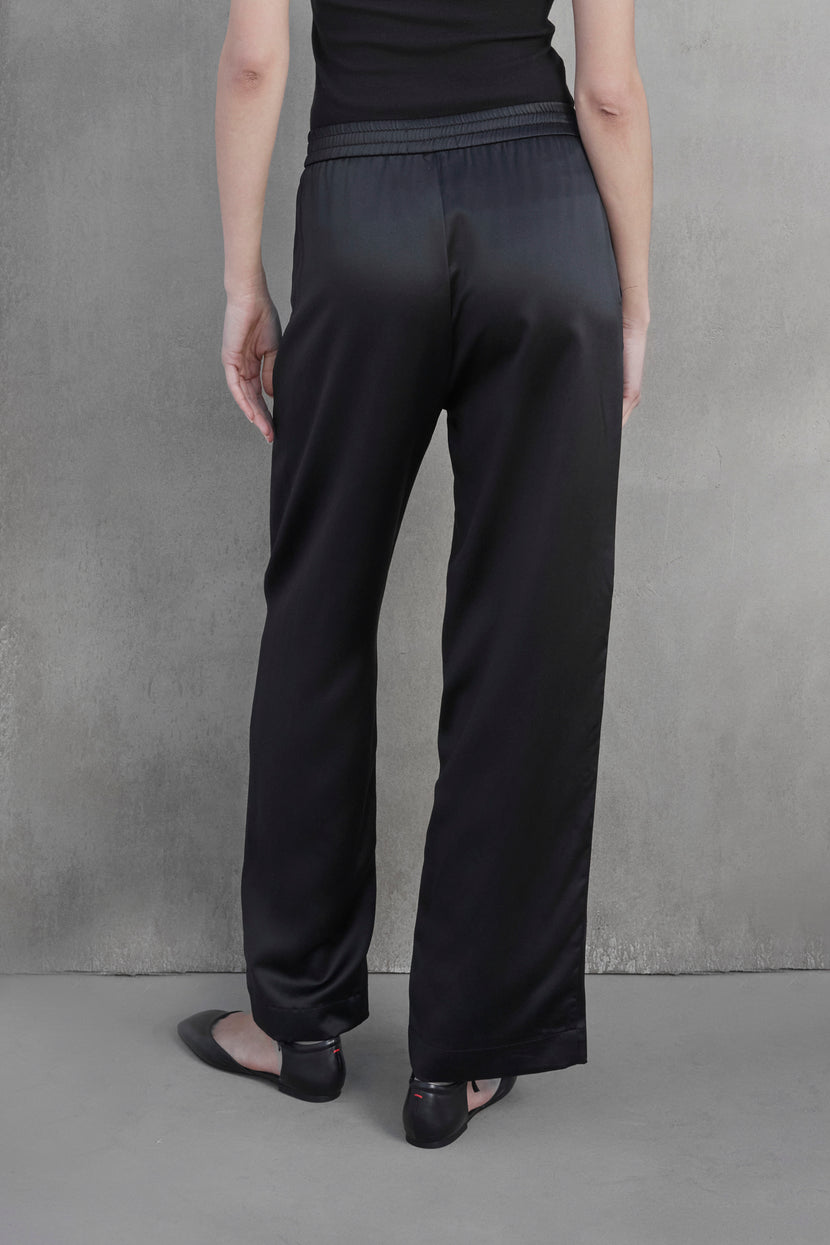 Person standing with their back to the camera, wearing black MODESTO SILK CHARMEUSE PANT by Velvet by Jenny Graham and black flats against a plain, light-colored wall.