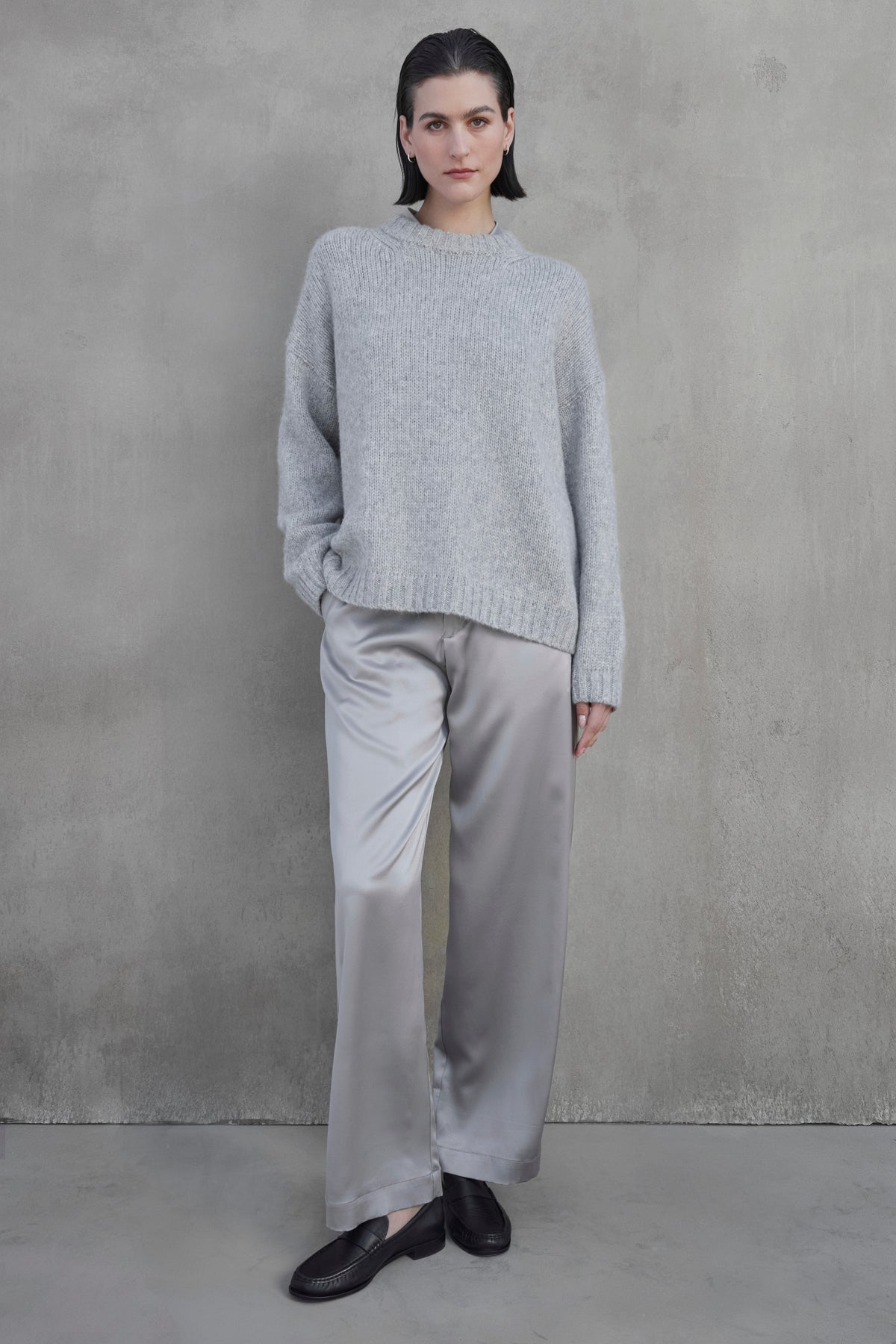 A person stands against a gray wall, wearing a gray CAMBRIA ALPACA SWEATER from Velvet by Jenny Graham, silver satin pants, and black loafers. Their left hand is in their pocket.-37659958640833