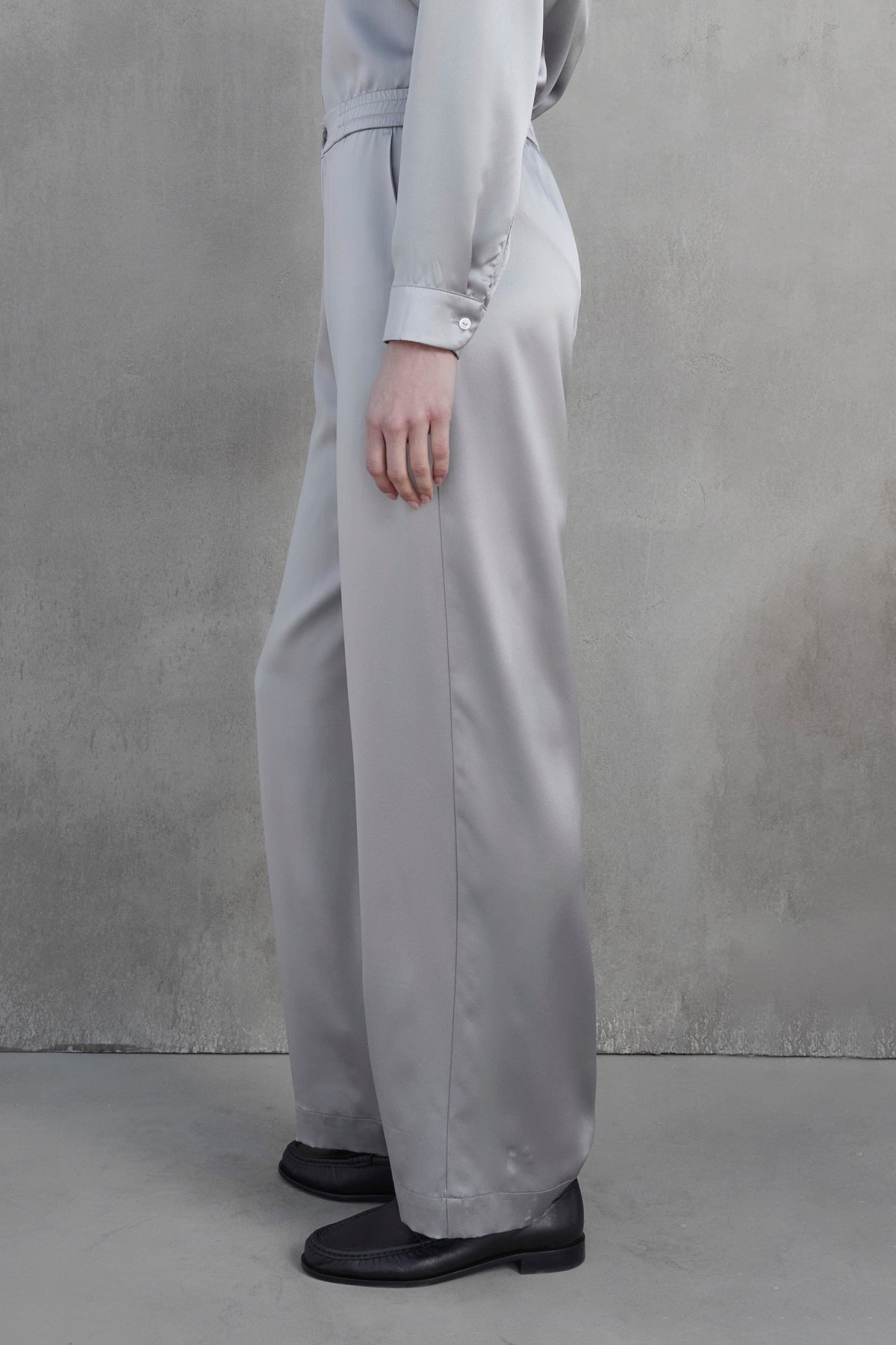   A person is standing sideways against a gray wall, wearing a light gray, buttoned long-sleeve shirt paired with the MODESTO SILK CHARMEUSE PANT by Velvet by Jenny Graham and black shoes. 