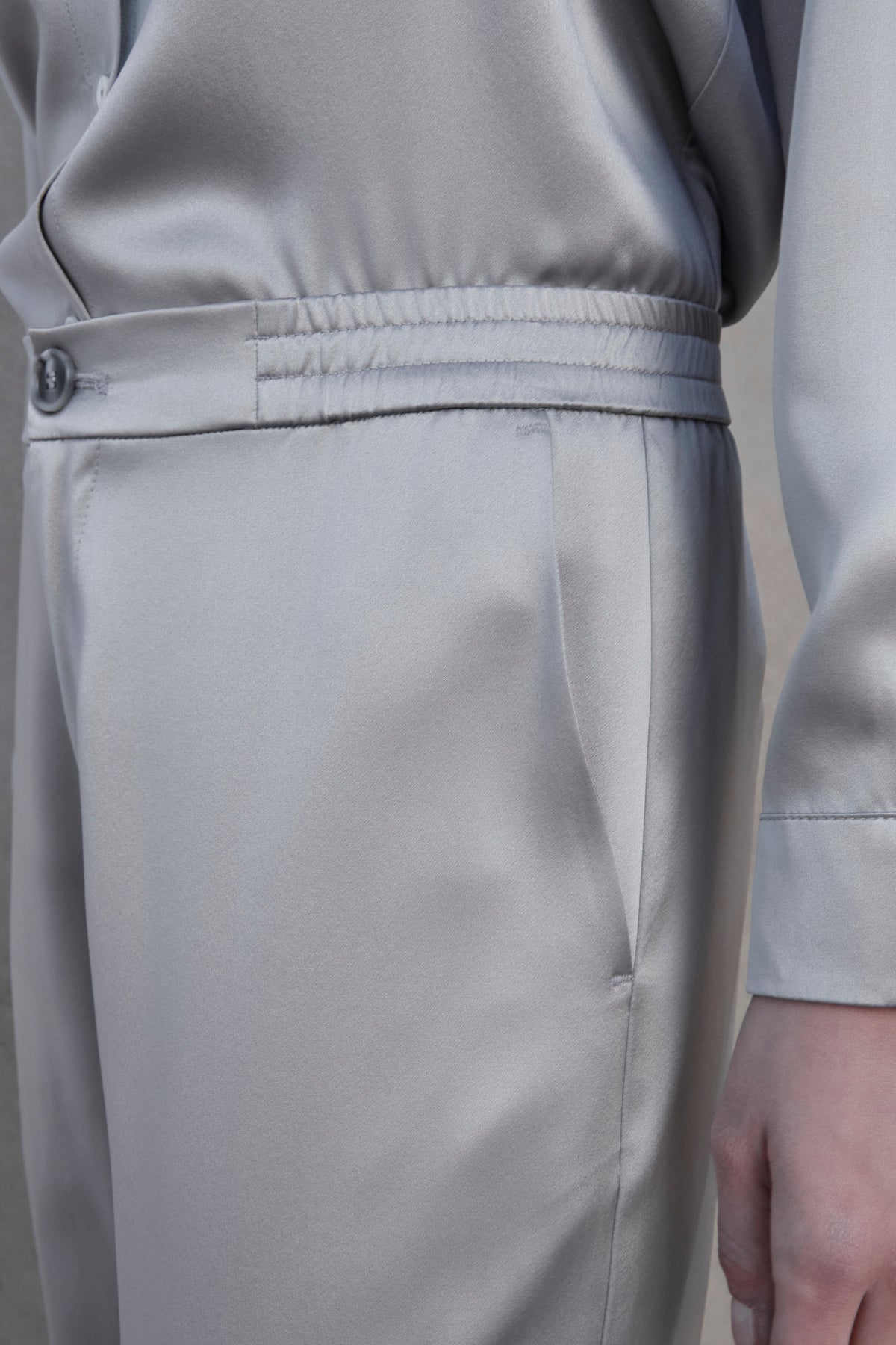   Close-up of a person wearing the MODESTO SILK CHARMEUSE PANT by Velvet by Jenny Graham, featuring grey silk wide-leg pants with an elastic waistband and button detail. Their hand rests by their side, and the fabric appears smooth and slightly reflective. 