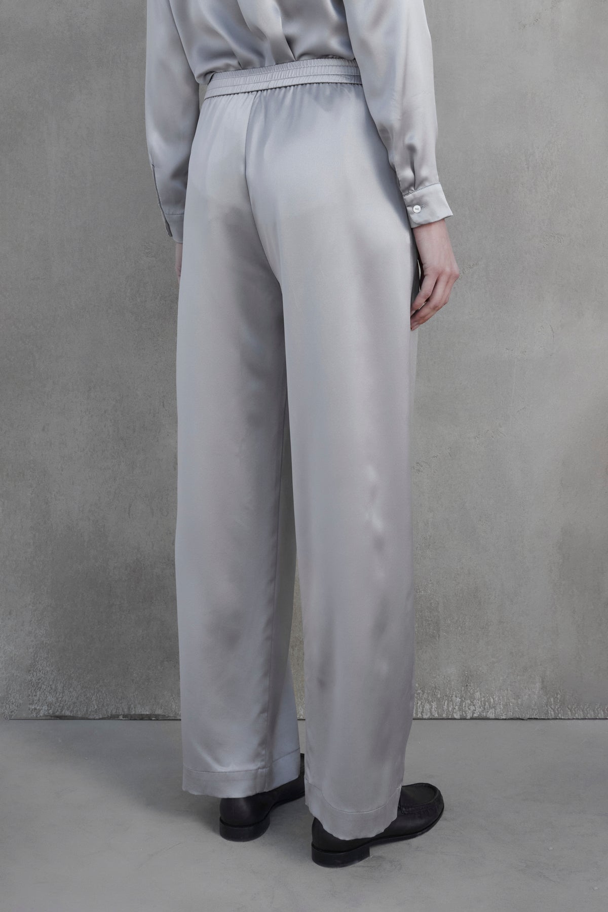   A person is shown from the back wearing Velvet by Jenny Graham's MODESTO SILK CHARMEUSE PANT, which are loose-fitting, high-waisted, and wide-legged, paired with a matching top featuring buttoned cuffs. The individual stands against a plain gray background. 