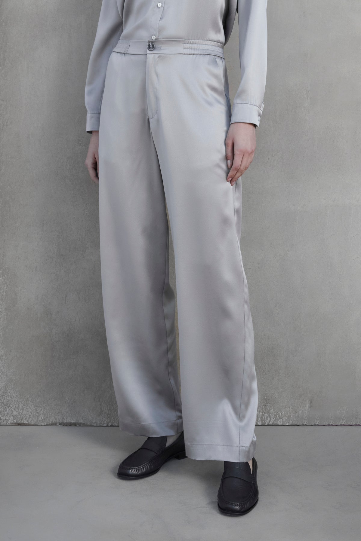   A person wearing a light gray shirt and matching MODESTO SILK CHARMEUSE PANT by Velvet by Jenny Graham, featuring an elastic waist and wide-leg design, stands against a plain gray background with hands relaxed by their sides. They are also wearing dark-colored shoes. 