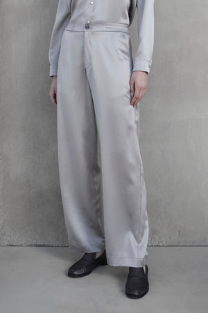 A person wearing a light gray shirt and matching MODESTO SILK CHARMEUSE PANT by Velvet by Jenny Graham, featuring an elastic waist and wide-leg design, stands against a plain gray background with hands relaxed by their sides. They are also wearing dark-colored shoes.