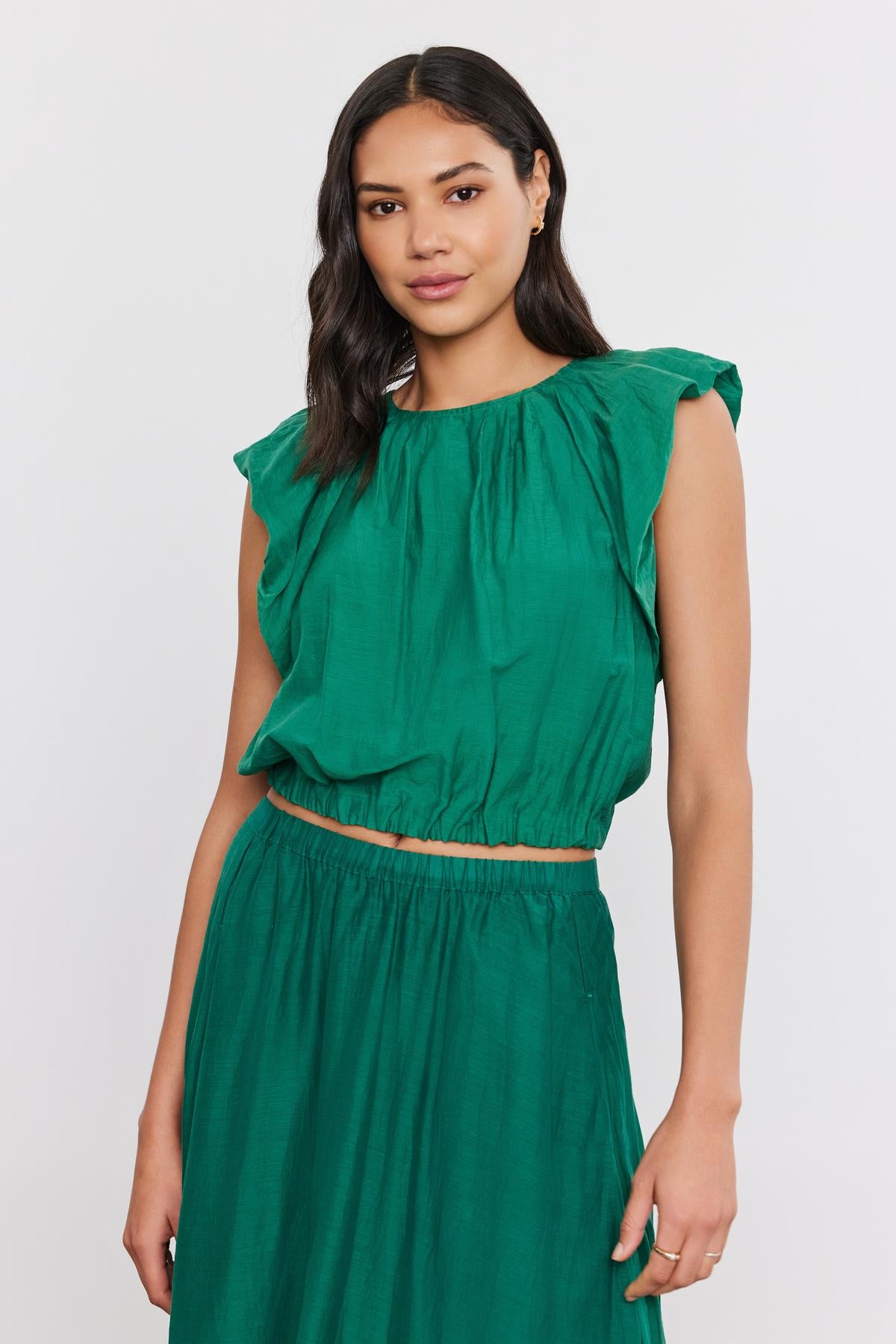   A woman in a green ruffled sleeveless AMORA TOP with a crew neckline and matching skirt, standing against a white background. (Brand: Velvet by Graham & Spencer) 