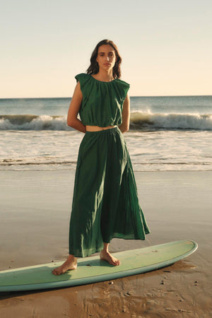 A woman in a green silk dress stands on a surfboard on the beach, with the ocean in the background, wearing an AMORA TOP from Velvet by Graham & Spencer.