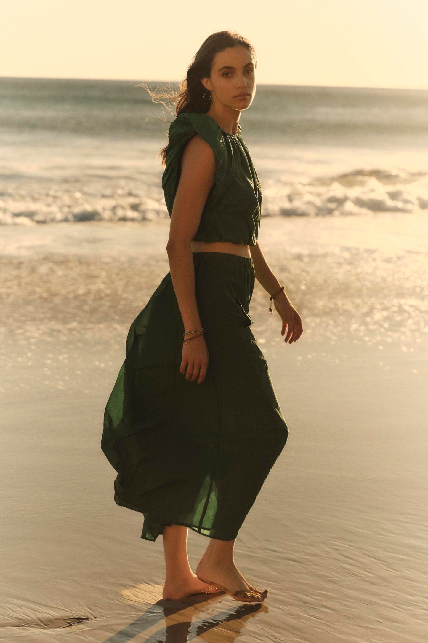 A woman in a flowing green Amora Top by Velvet by Graham & Spencer walks barefoot along a beach at sunset, looking over her shoulder.-36910240563393