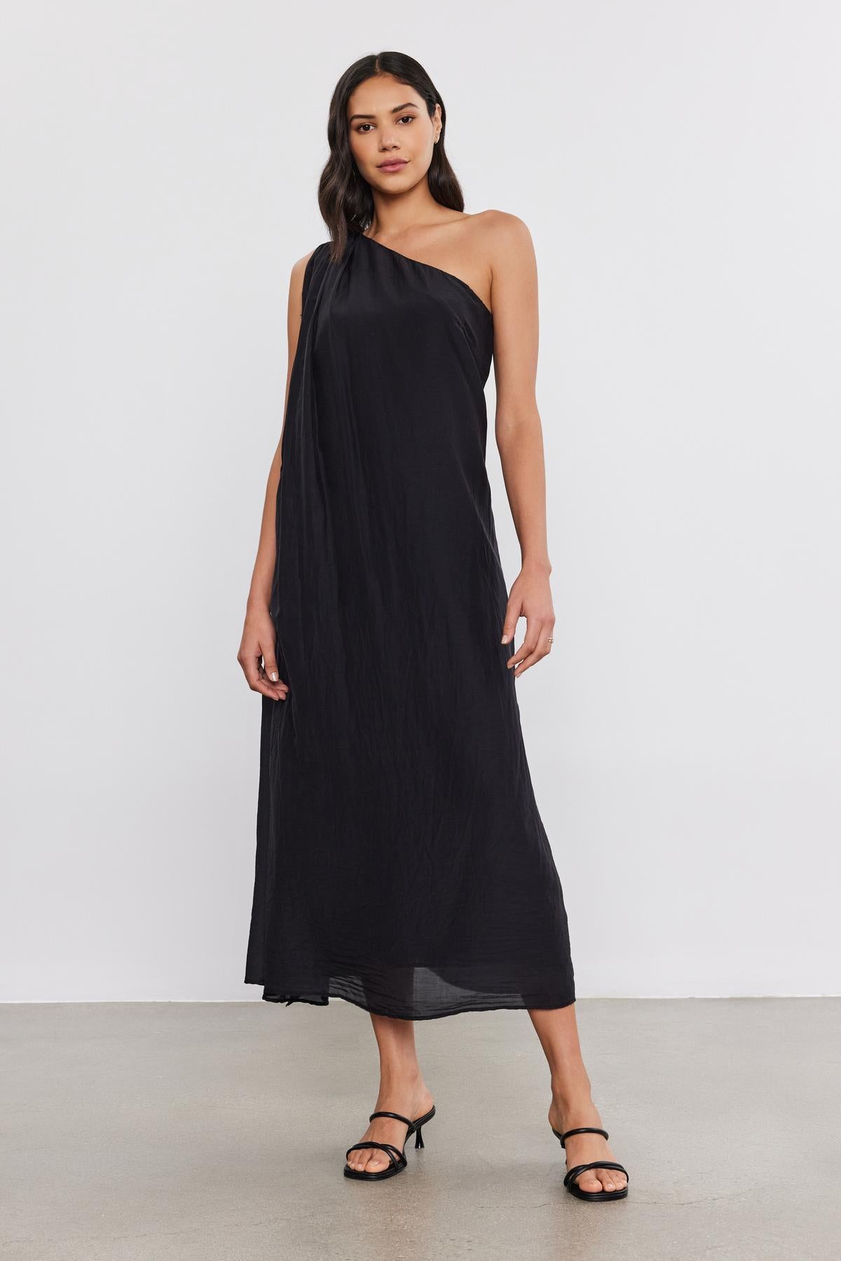   Woman in a black one-shoulder Velvet by Graham & Spencer DIANA DRESS and black heels, standing on a plain backdrop. 