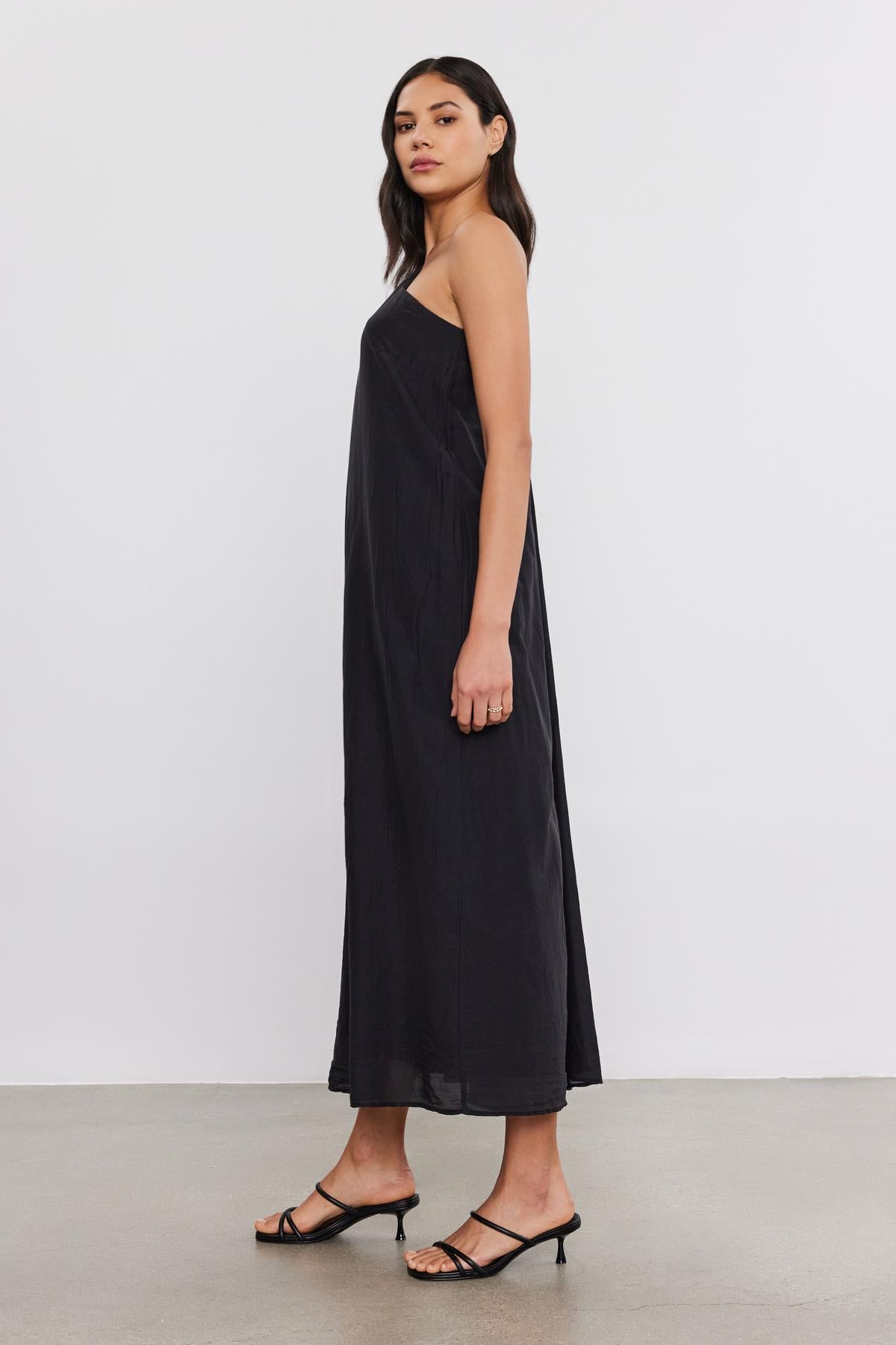   A woman in a black Velvet by Graham & Spencer DIANA DRESS and strappy sandals, standing side-on in a studio with a grey background. 