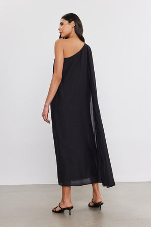 A woman stands with her back to the camera, wearing a flowing black one-shoulder Diana Dress made of silk cotton voile by Velvet by Graham & Spencer and black high heels.