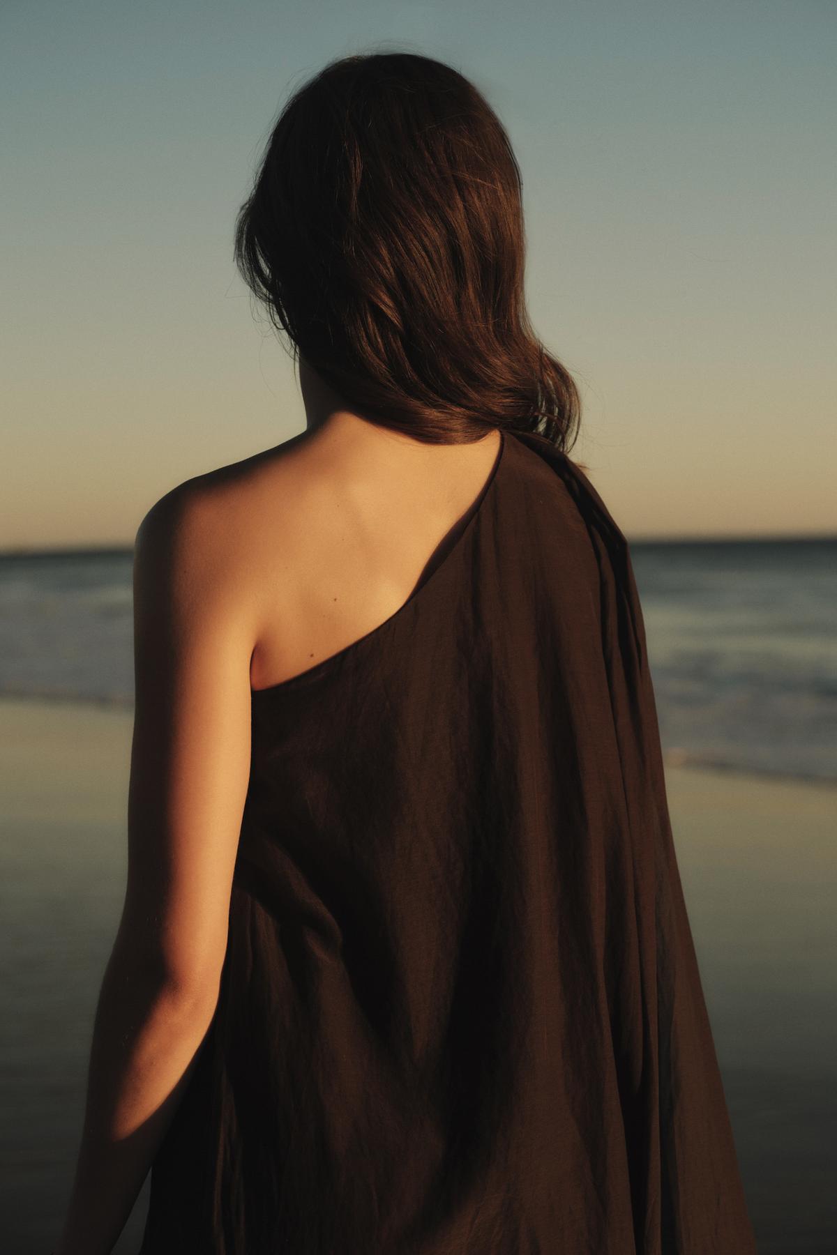 A woman draped in a Velvet by Graham & Spencer DIANA DRESS stands facing the ocean at sunset, her back to the camera.-36918818996417