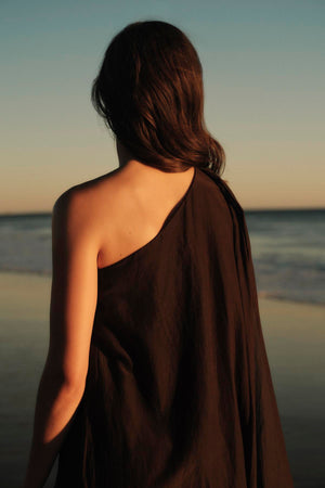 A woman draped in a Velvet by Graham & Spencer DIANA DRESS stands facing the ocean at sunset, her back to the camera.