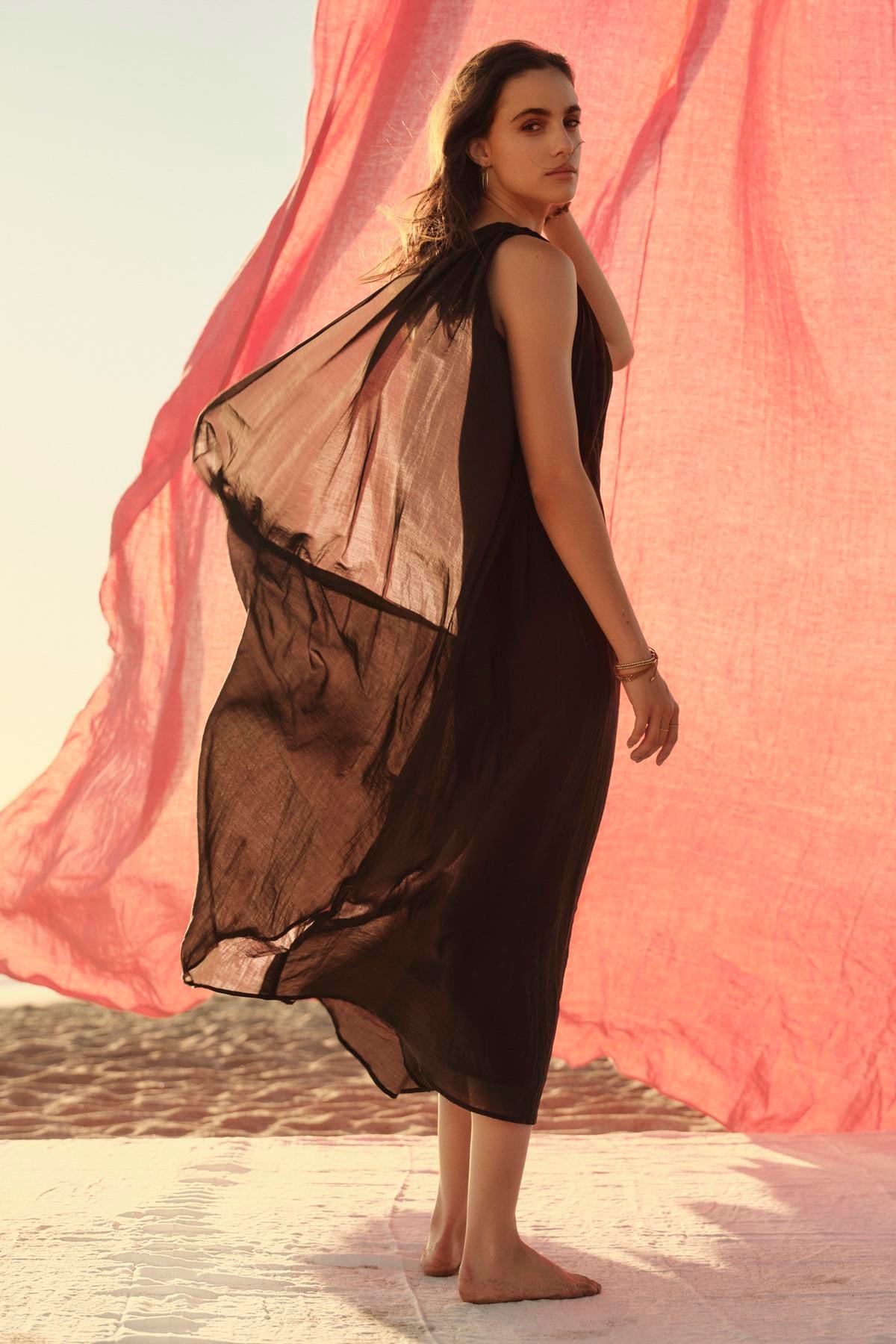   A woman in a flowing one-shoulder black Velvet by Graham & Spencer Diana dress stands barefoot, looking back, with a large pink fabric billowing in the background against a soft-lit sky. 