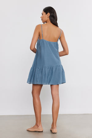 A person stands facing away from the camera, wearing a blue sleeveless mini length SUZIE DRESS by Velvet by Graham & Spencer with a ruffled hem and tan flat shoes, against a plain white background.