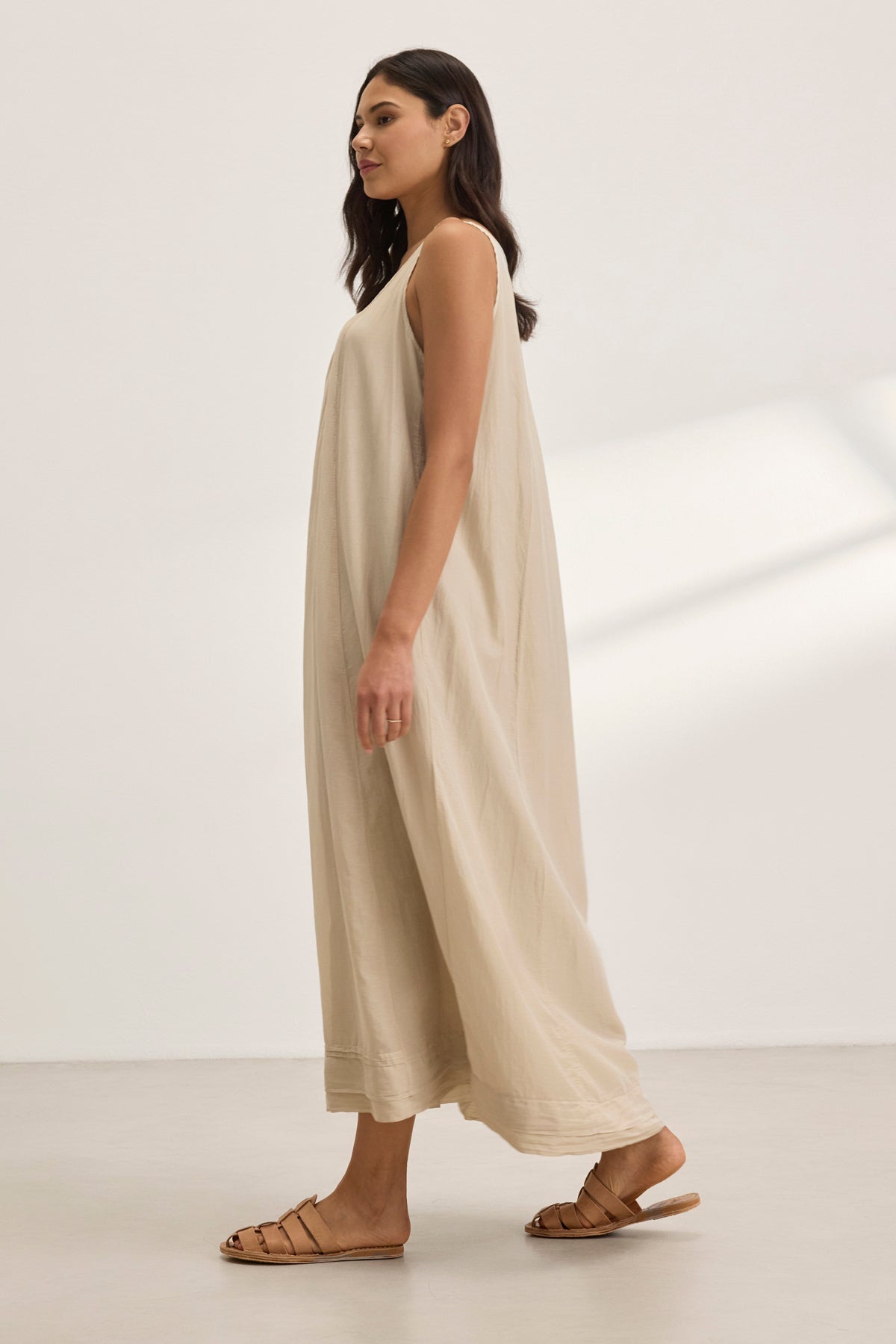   A woman in a BAYOU SILK COTTON VOILE DRESS by Velvet by Graham & Spencer, featuring a scoop neckline, stands against a plain, light background. The elegant beige maxi dress gracefully drapes down to her ankles, paired with brown sandals. 