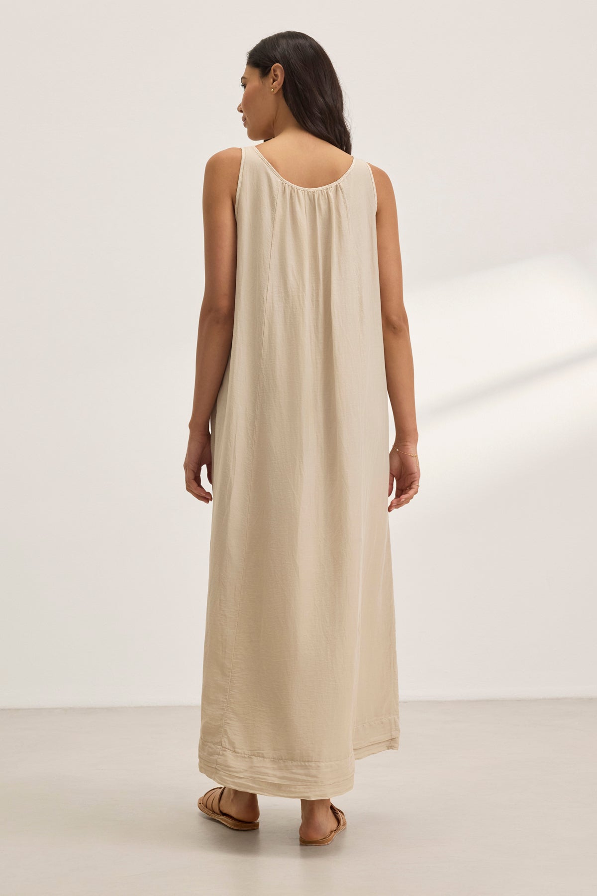   Someone in the BAYOU SILK COTTON VOILE DRESS by Velvet by Graham & Spencer, a sleeveless floor-length beige dress, stands on a light-colored floor facing away. The silk cotton voile fabric adds elegance for versatile styling in any setting. 