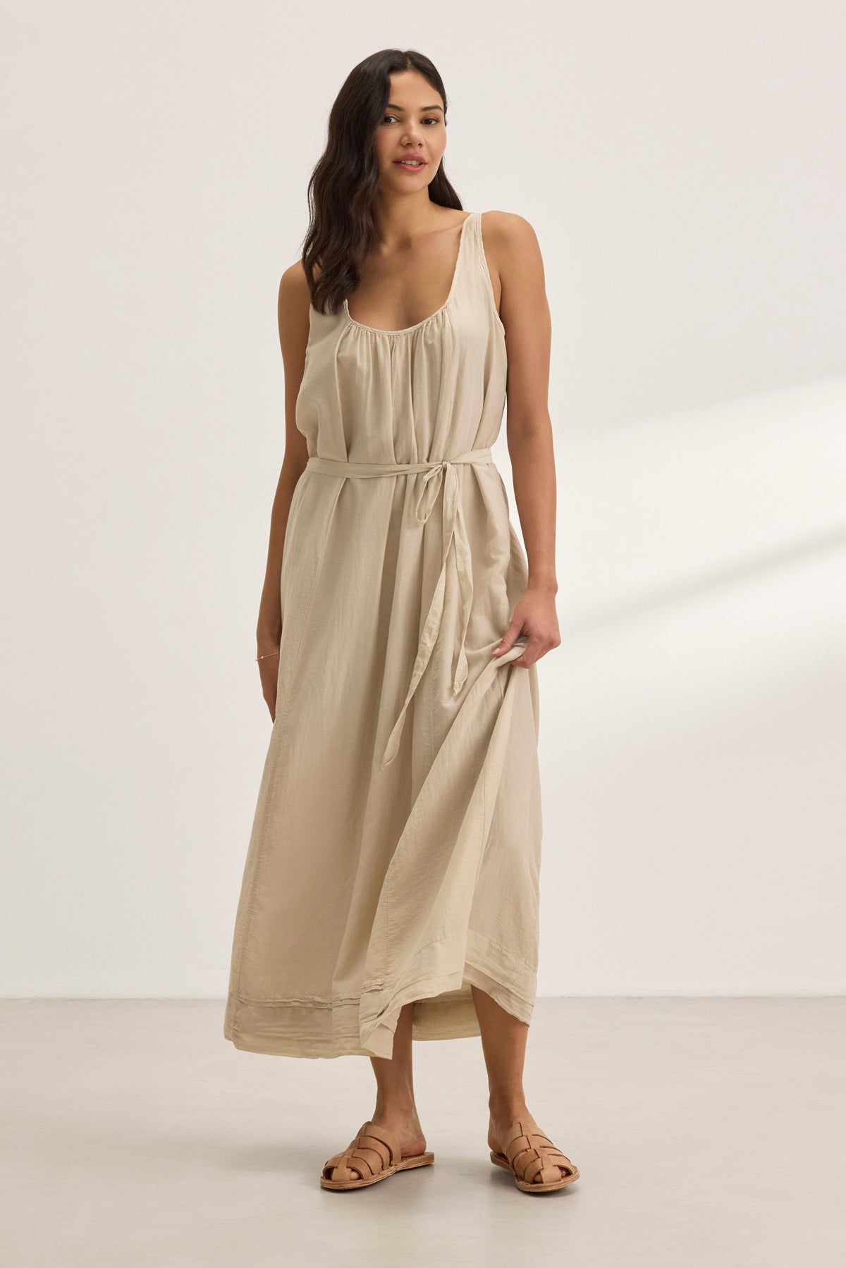 In a neutral-toned room, a woman elegantly wears the BAYOU SILK COTTON VOILE DRESS by Velvet by Graham & Spencer, a sleeveless beige maxi dress made from luxurious silk cotton voile, showcasing its versatile styling.-38801131208897