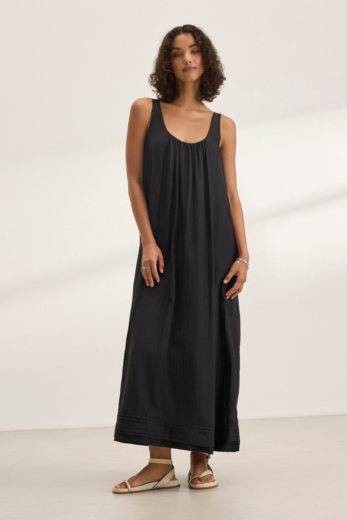   A woman showcases the BAYOU SILK COTTON VOILE DRESS by Velvet by Graham & Spencer, a sleek sleeveless black maxi, paired with sandals. She stands on a light-colored floor against a white background, highlighting its versatile styling perfect for any occasion. 