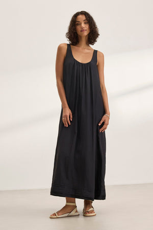 A woman showcases the BAYOU SILK COTTON VOILE DRESS by Velvet by Graham & Spencer, a sleek sleeveless black maxi, paired with sandals. She stands on a light-colored floor against a white background, highlighting its versatile styling perfect for any occasion.