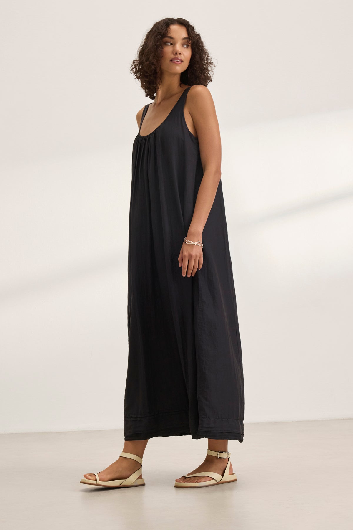   In a neutral backdrop, a person wearing the BAYOU SILK COTTON VOILE DRESS by Velvet by Graham & Spencer gazes slightly left. The sleeveless, long black maxi dress elegantly drapes, paired with sandals and a bracelet for versatile styling. 