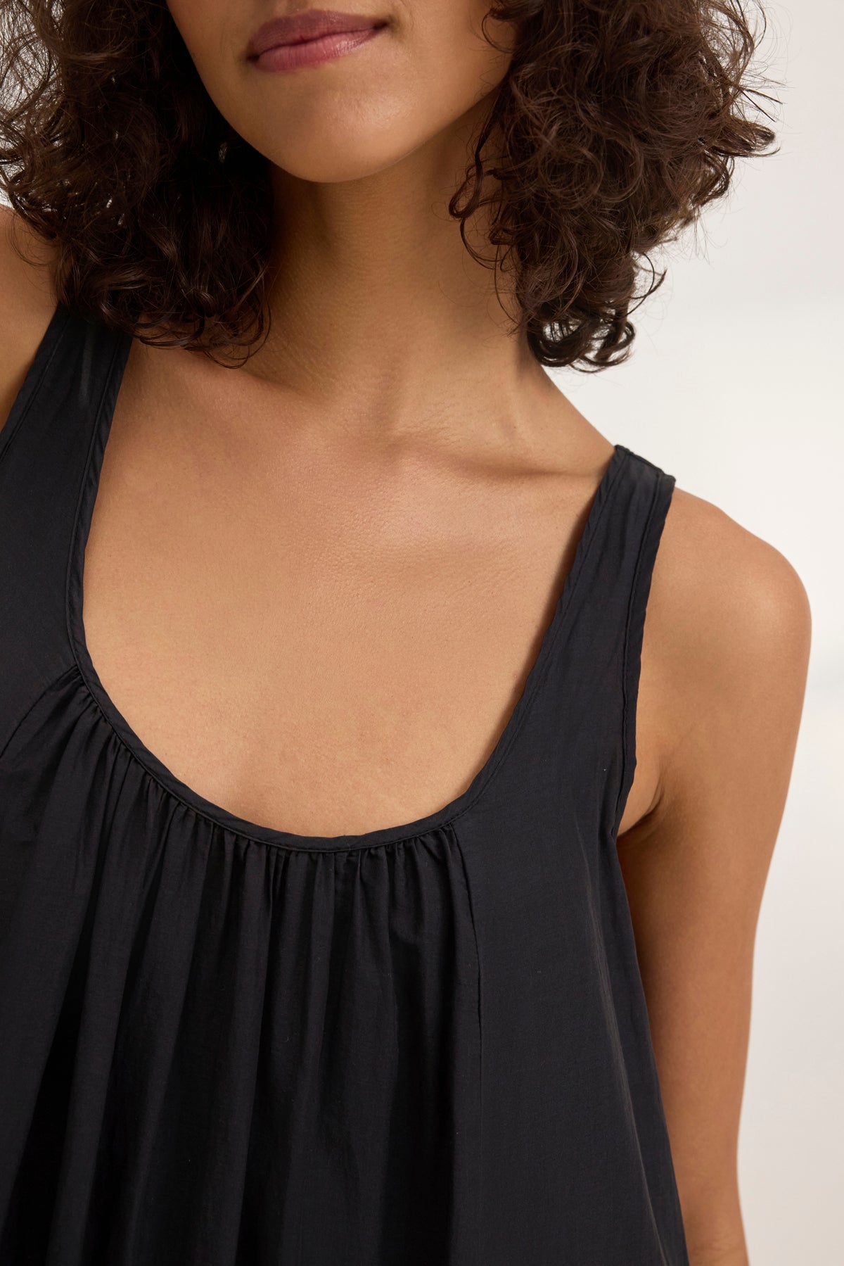   A curly-haired person in a sleeveless black top showcases versatile styling that pairs effortlessly with the BAYOU SILK COTTON VOILE DRESS by Velvet by Graham & Spencer. The close-up captures their upper torso and part of the face. 