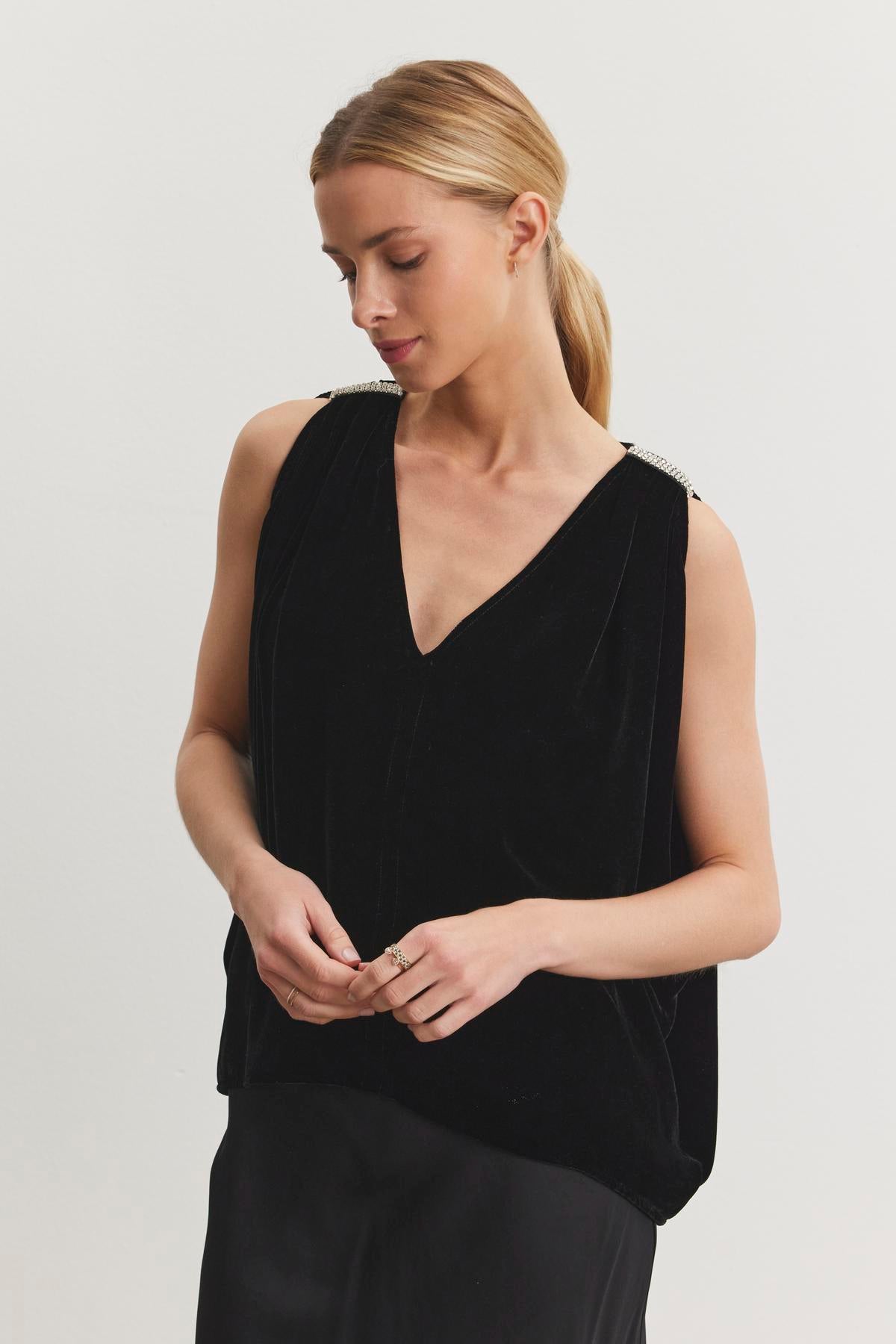   A person dressed in the ADELLE SILK VELVET TOP by Velvet by Graham & Spencer, along with a matching black skirt, stands against a plain background. They gaze downward with hands lightly clasped, exuding the elegance reminiscent of holiday gatherings. 