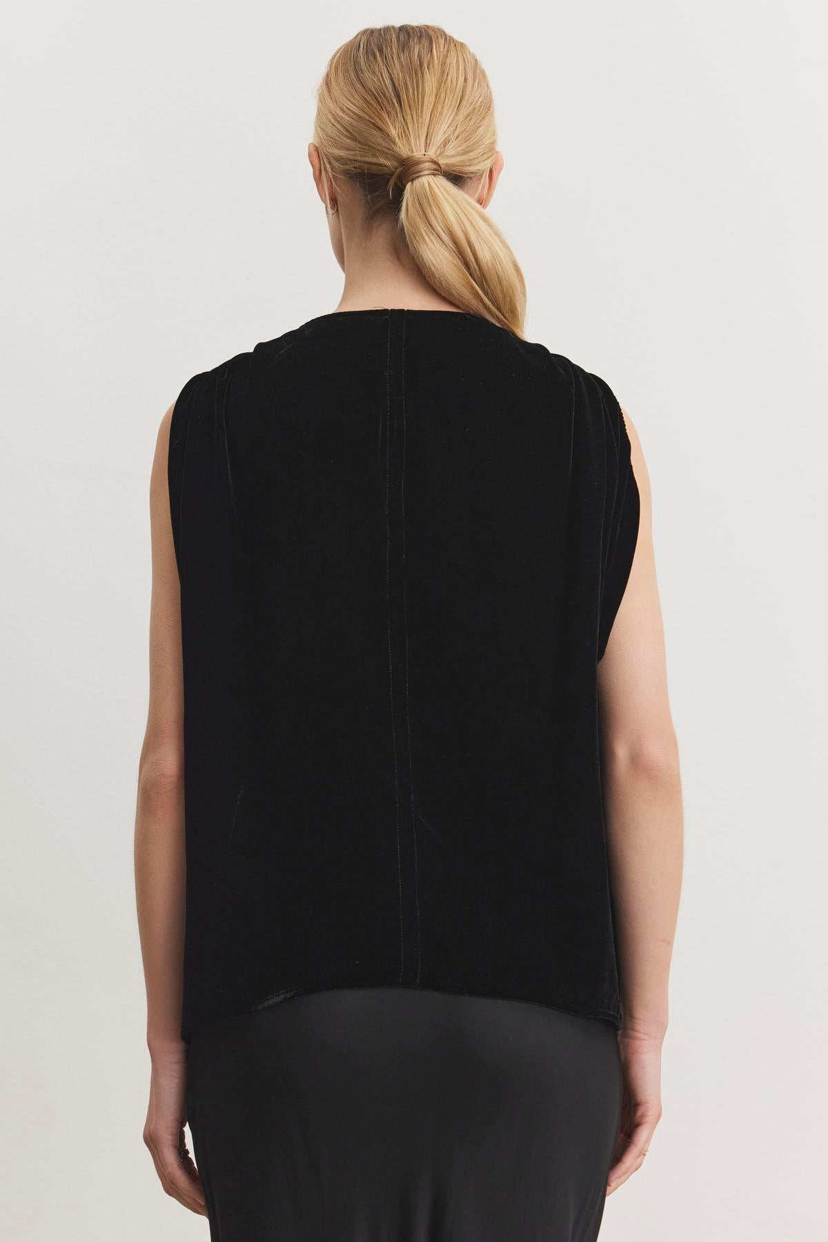   The rear view showcases an individual with blond hair styled in a low ponytail, dressed in the ADELLE SILK VELVET TOP by Velvet by Graham & Spencer over a black skirt, positioned against a plain white background. Ideal for holiday gatherings. 