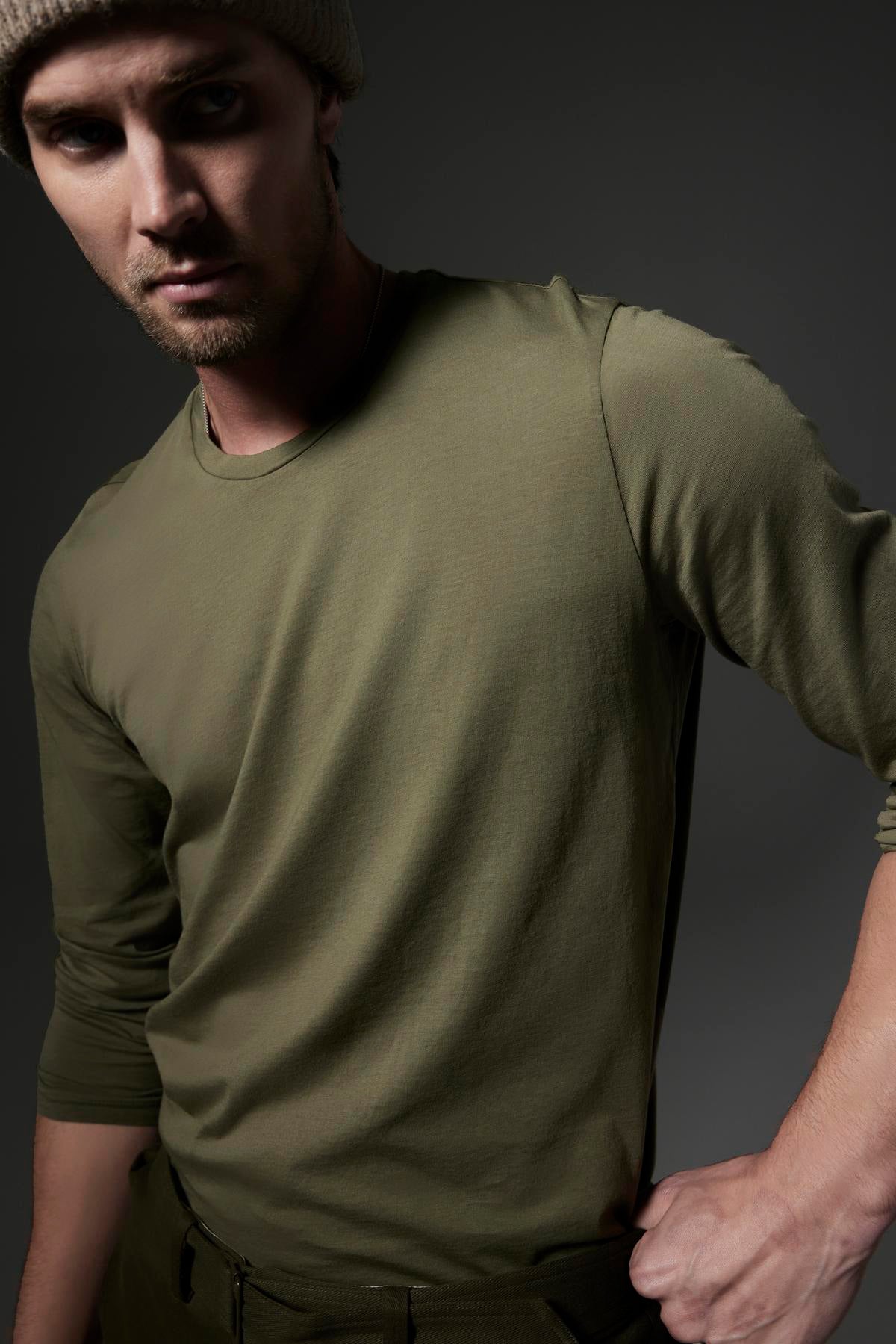   A man with an intense expression, wearing a versatile SKEETER TEE long-sleeve shirt by Velvet by Graham & Spencer and a knit beanie, poses against a dark background. 