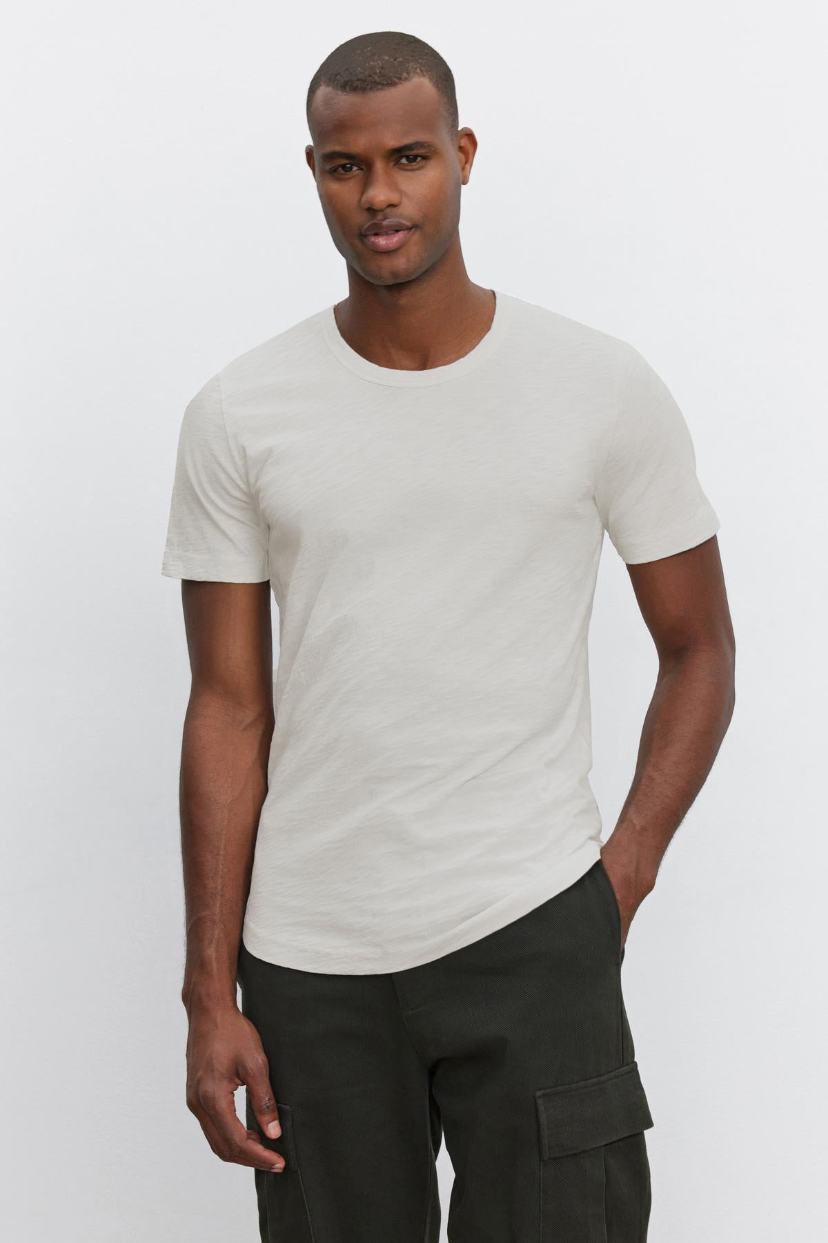   A man standing against a plain white background wears the AMARO TEE, a light gray t-shirt with a classic crew neckline from Velvet by Graham & Spencer, paired with dark green cargo pants. 