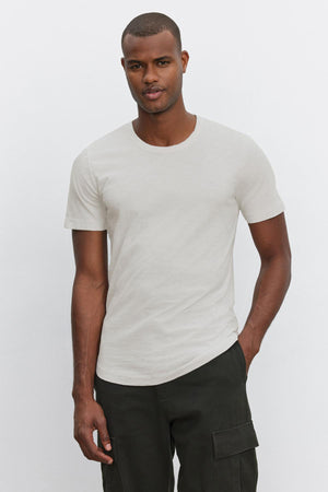 A man standing against a plain white background wears the AMARO TEE, a light gray t-shirt with a classic crew neckline from Velvet by Graham & Spencer, paired with dark green cargo pants.