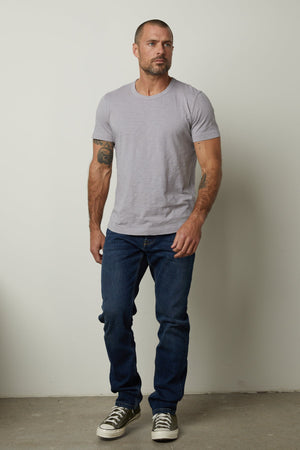 A man with short hair and tattoos on his arms stands against a plain wall, wearing a light gray, relaxed fit Velvet by Graham & Spencer AMARO TEE, blue jeans, and gray sneakers.