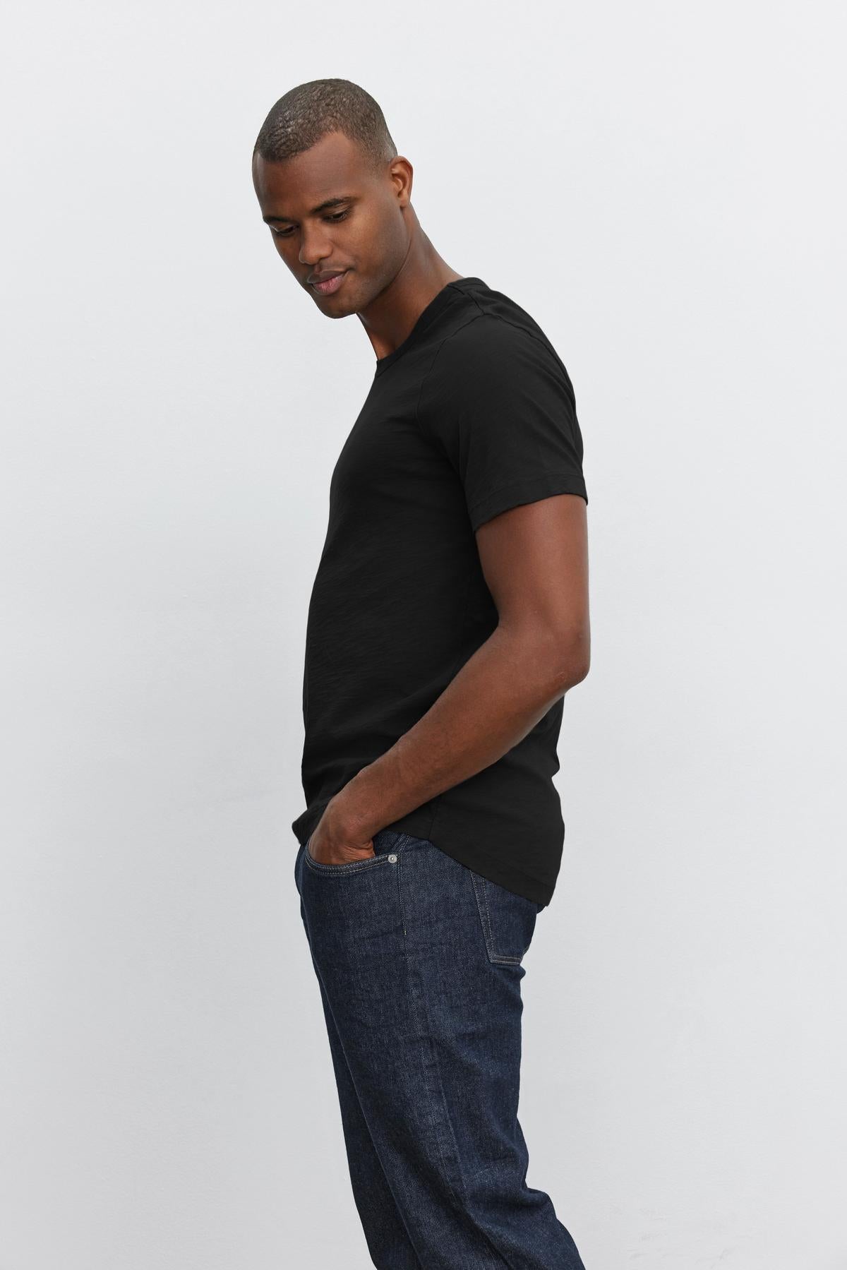   Man in a black crew neck AMARO TEE with heathered texture and jeans standing sideways against a white background by Velvet by Graham & Spencer. 